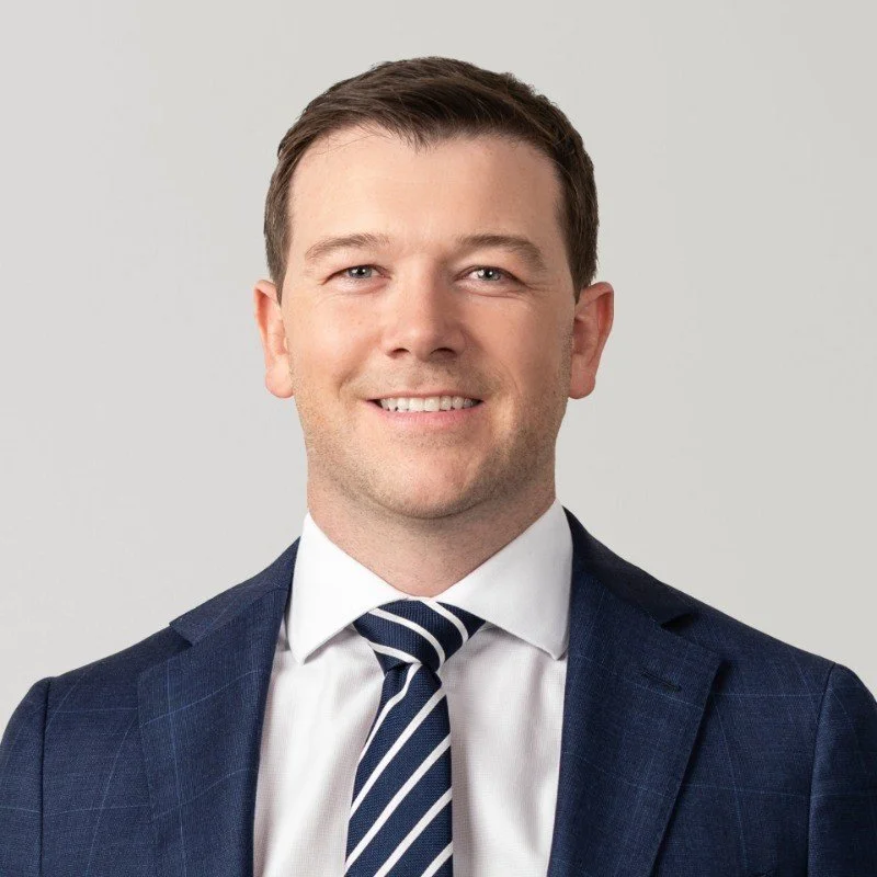 A professional headshot of a man in a navy blue suit, white shirt, and diagonally striped navy and white tie, smiling against a plain light gray background.
