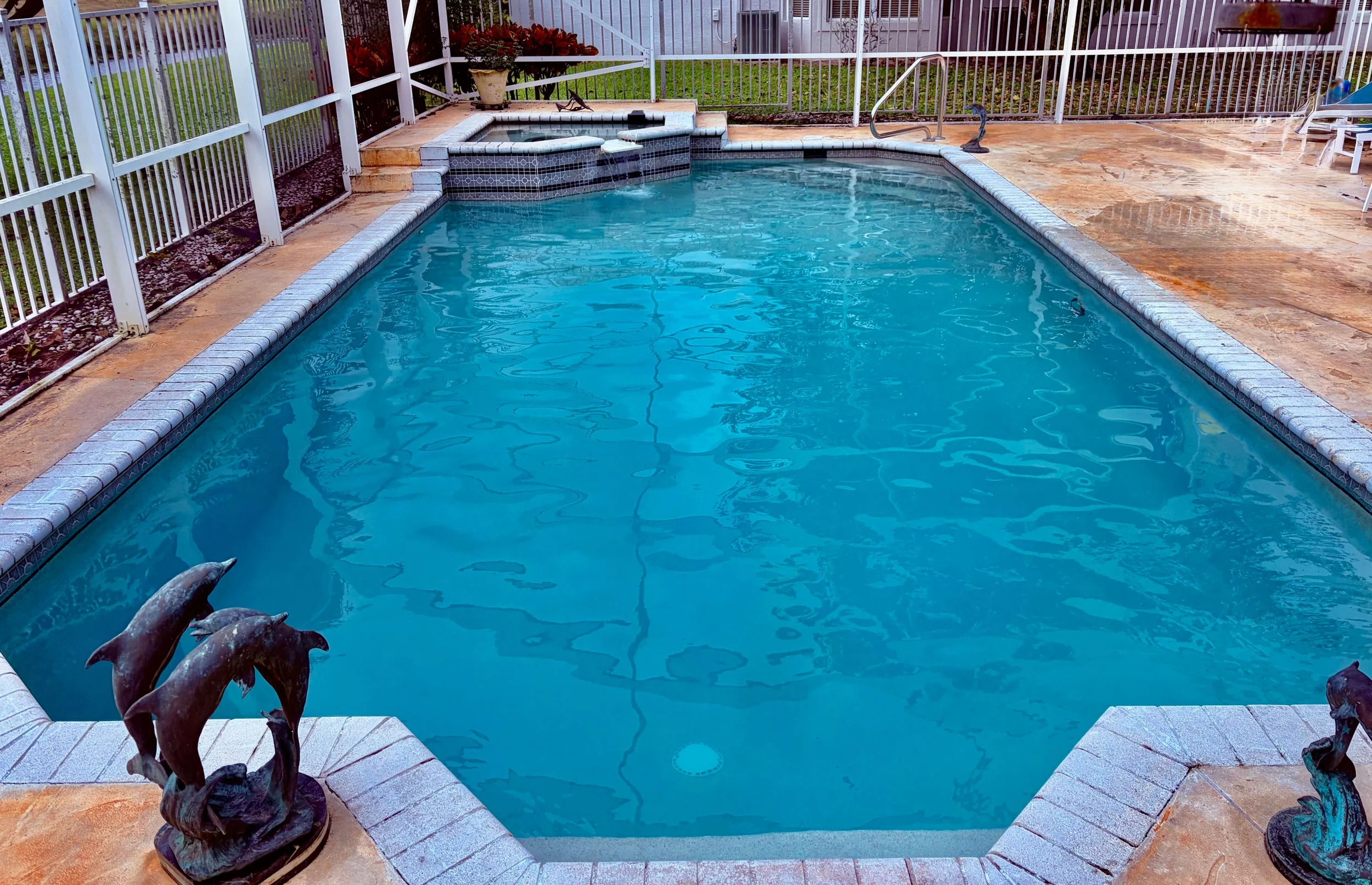 A backyard swimming pool with dolphin statues on the corners, enclosed by a white metal fence, surrounded by a stone deck and some potted plants.