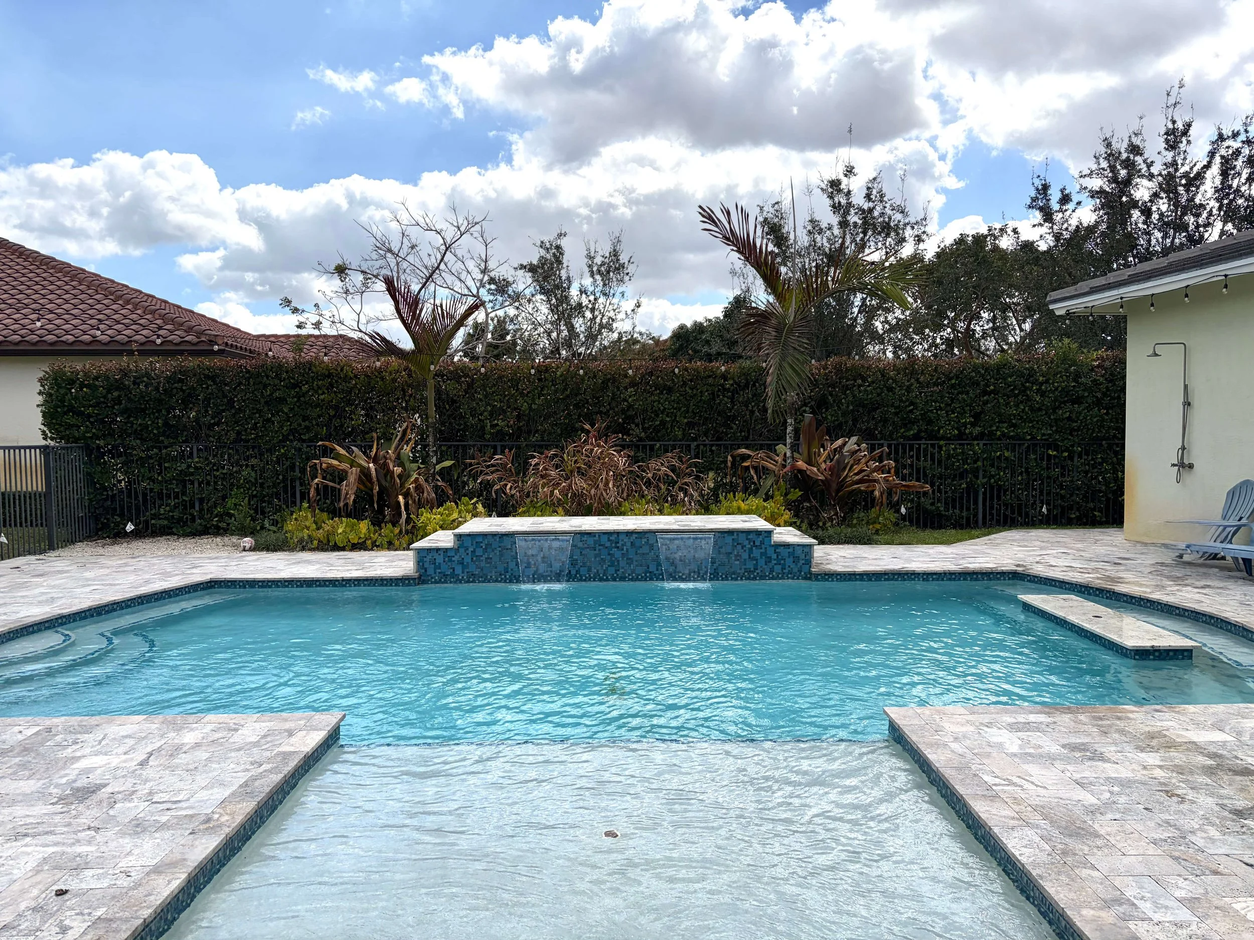 Residential backyard with a swimming pool and spa, surrounded by stone paving, lush plants, palm trees, and a hedge, with a house and cloudy sky in the background.