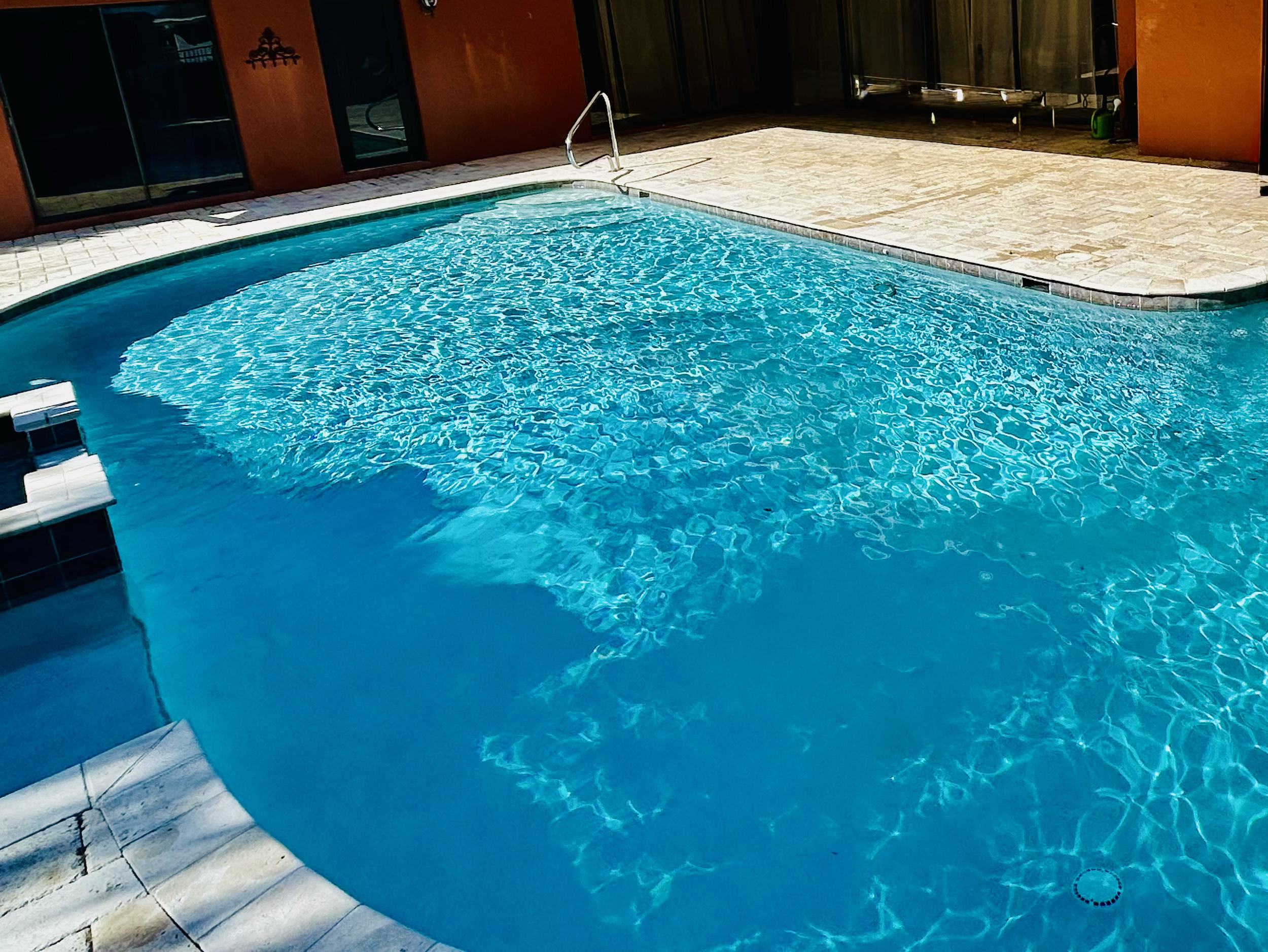 A swimming pool with clear blue water outside a building, with steps leading into the water and a metal handrail at the pool's edge.
