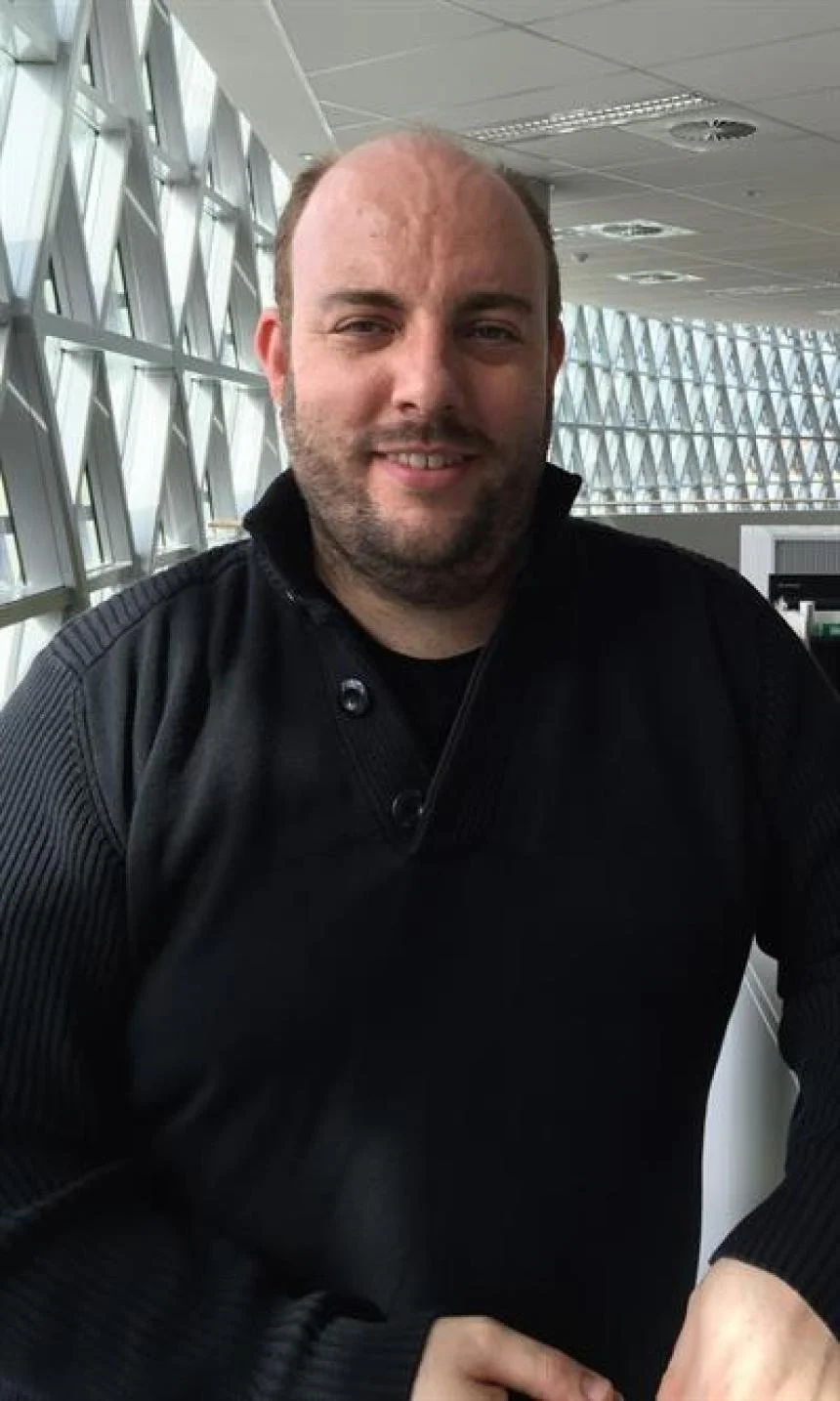 A smiling man with a beard and bald head sitting in an airport terminal with large windows behind him.