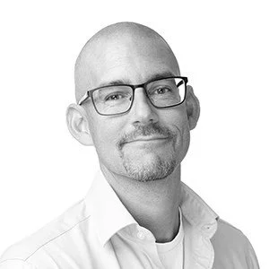 A black and white portrait of a man with glasses, a goatee, and a shaved head, wearing a collared shirt with a confident expression.