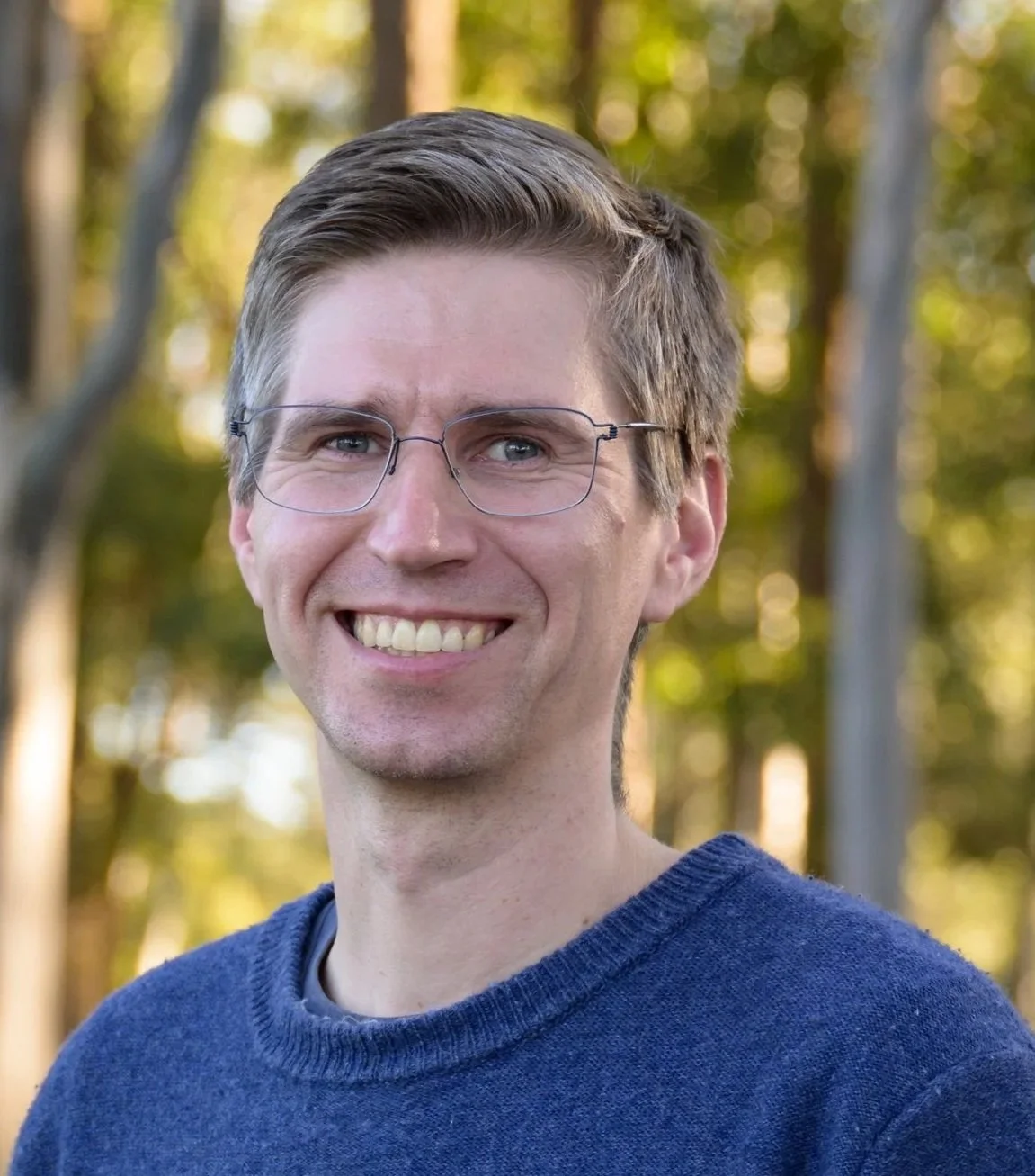 A smiling man with light skin, glasses, and short brown hair wearing a blue sweater, standing outdoors with trees in the background.