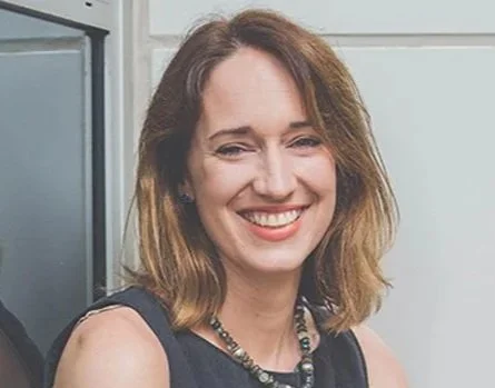 A smiling woman with shoulder-length brown hair wearing a black sleeveless top and necklace, standing indoors.