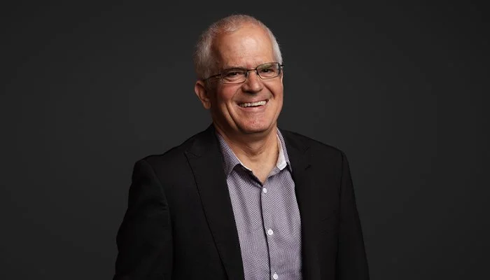 A smiling older man with glasses wearing a black blazer and a light-colored, patterned shirt posing against a dark background.