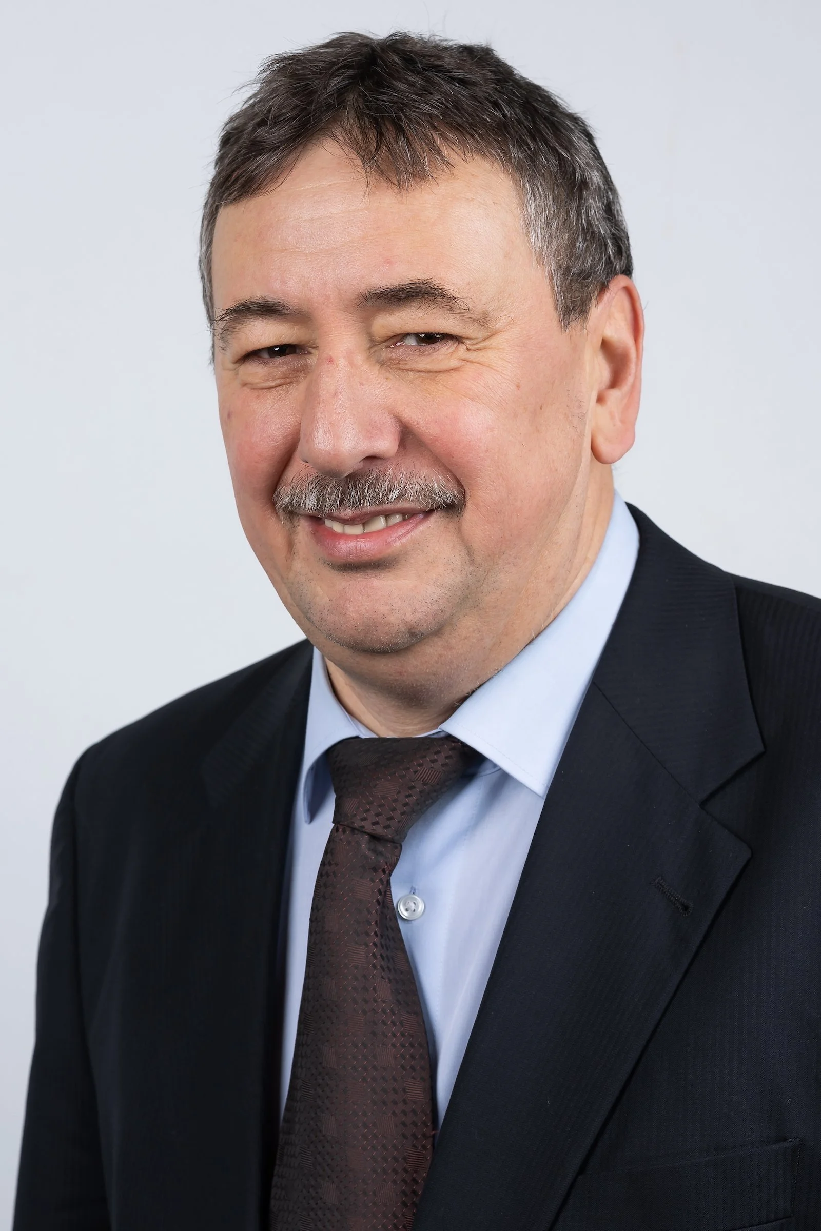 Portrait of a middle-aged man with brown hair, mustache, wearing a black suit, white shirt, and black tie, against a plain gray background.