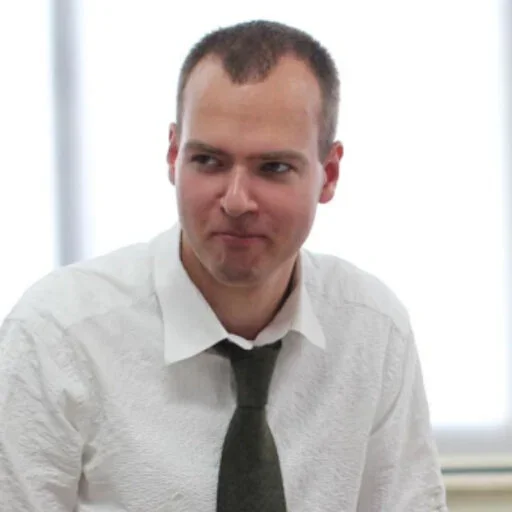 A young man with short brown hair, wearing a white dress shirt and a dark tie, sitting indoors with a neutral expression.