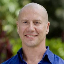 Close-up of a smiling man with a shaved head wearing a blue shirt outdoors