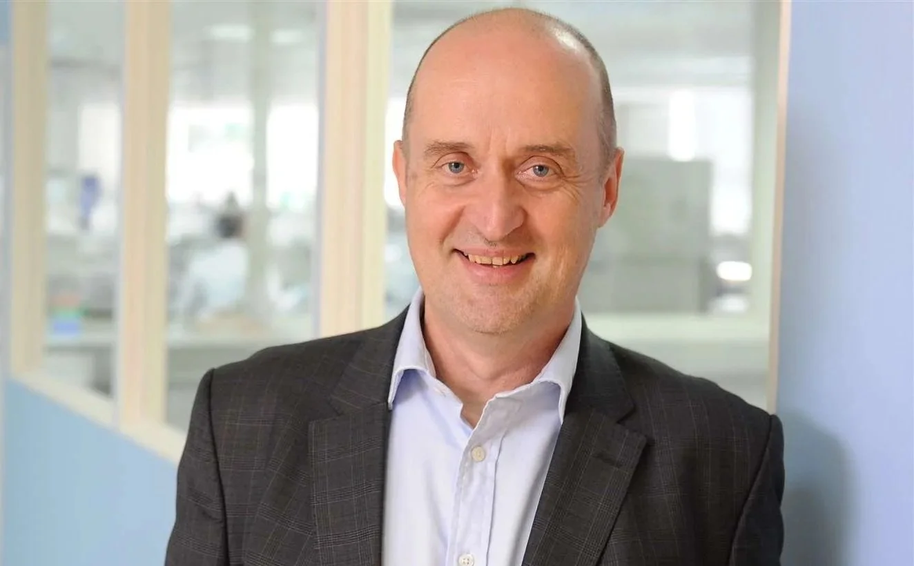 Portrait of a smiling middle-aged man with a bald head, wearing a dark plaid blazer and white collared shirt, standing in an office corridor with glass-walled offices in the background.