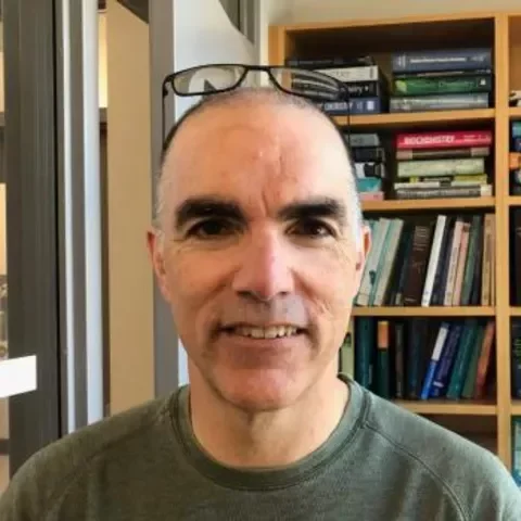 A man with glasses on his head smiling for the camera inside a room with bookshelves filled with books.