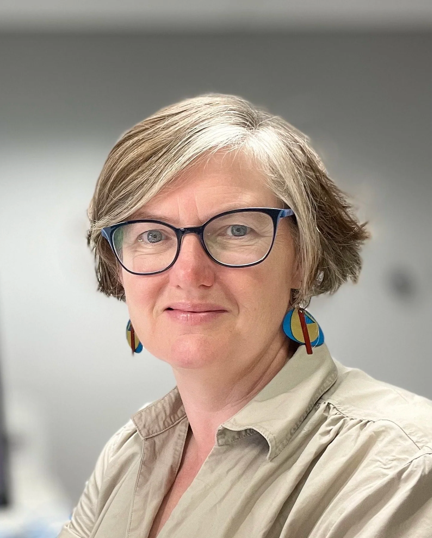 A woman with short, gray hair wearing glasses, colorful earrings, and a beige shirt, smiling in an indoor setting with a plain gray background.