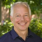 Portrait of a man with short gray hair, smiling, wearing a dark blue collared shirt, outdoors with greenery in the background.