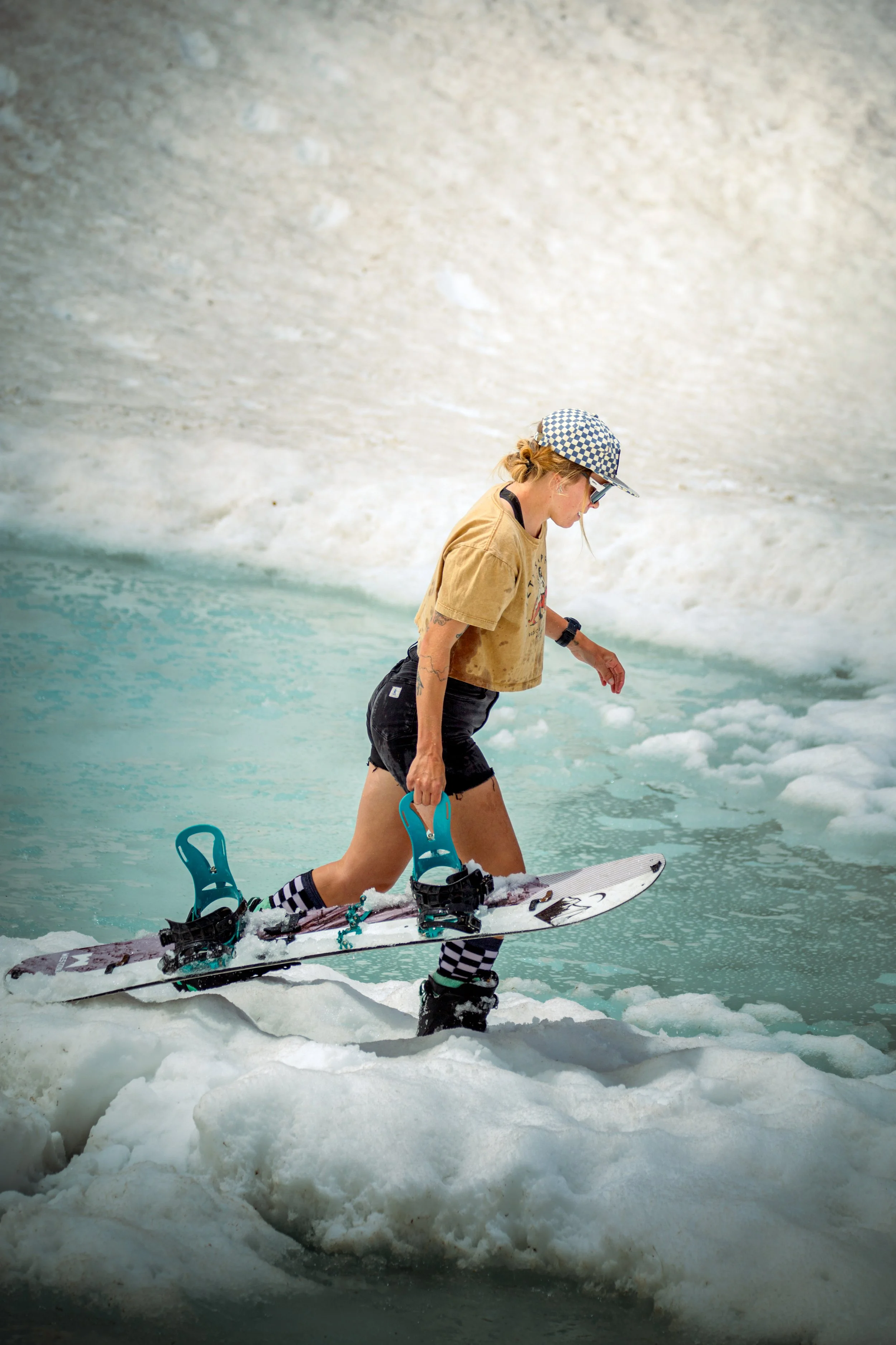 A woman wearing a beige t-shirt, black shorts, striped socks, and a checkered hat snowboarding on a snowy surface near a body of water.