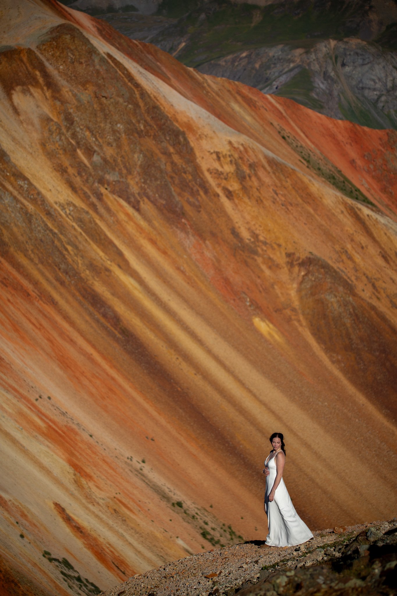 A woman in a white dress standing on rocky ground in front of colorful mountain slopes with a diagonal perspective.