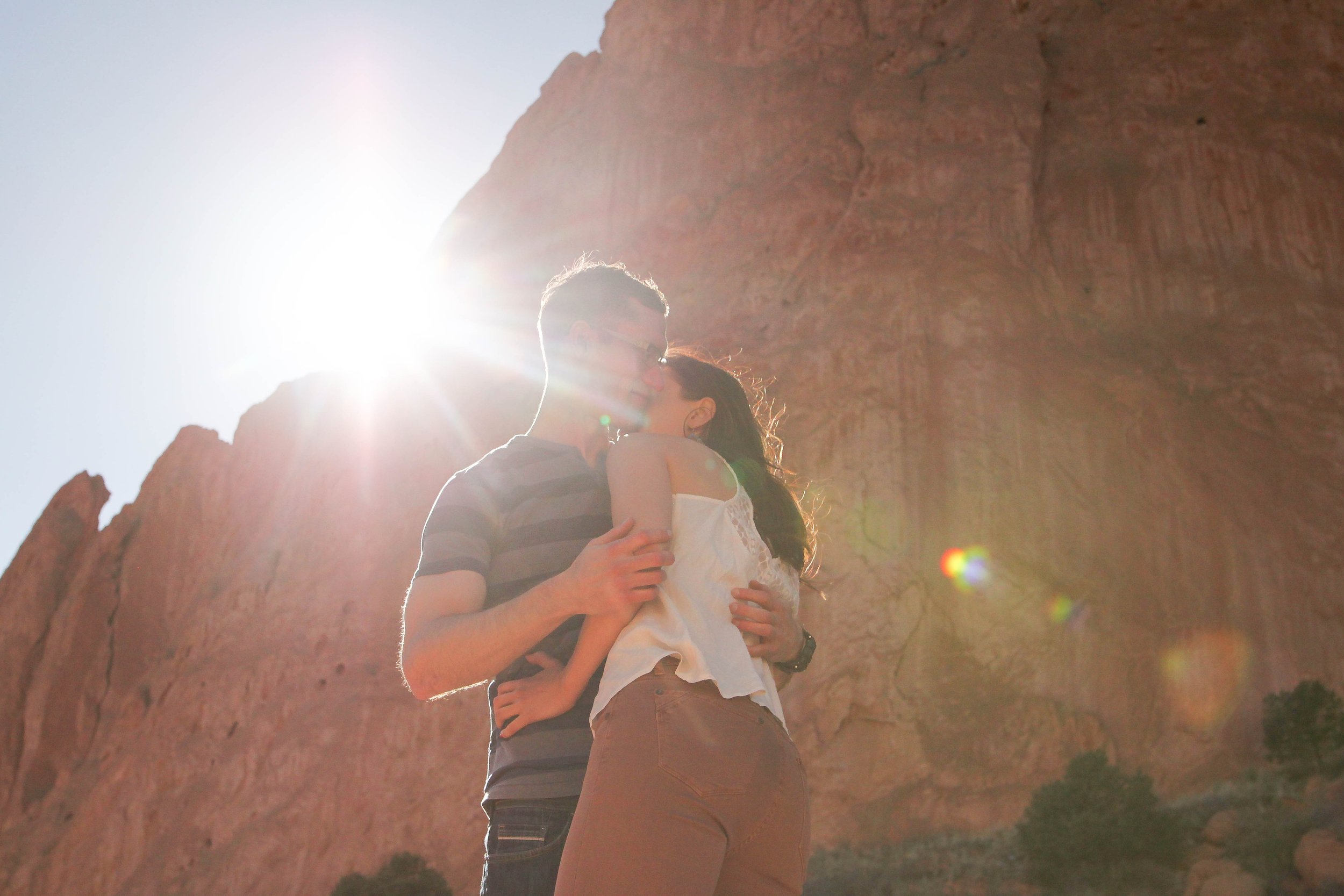 A couple kissing outdoors in front of a large rock formation with the sun shining brightly behind them.
