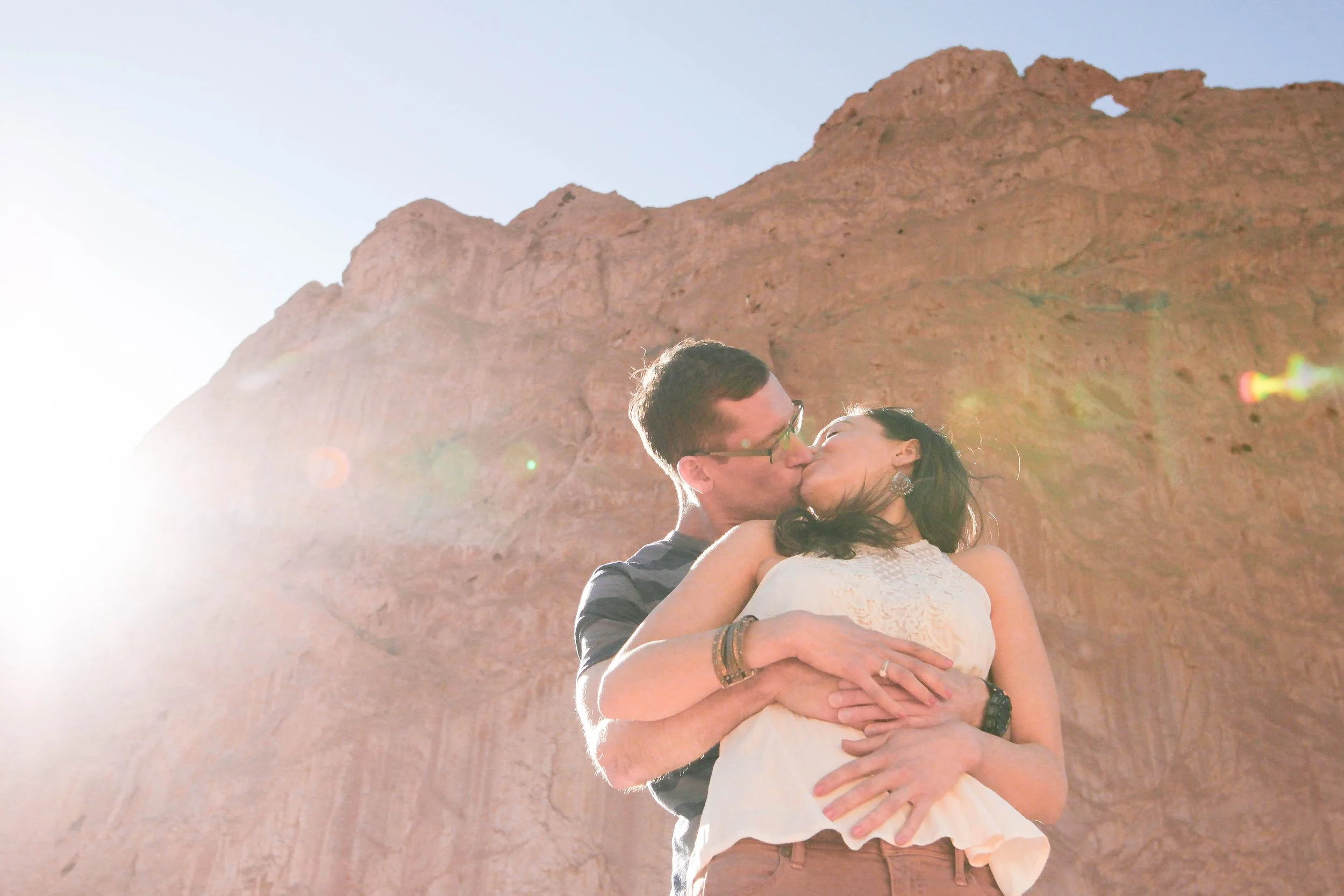 A couple kissing in front of a rocky desert landscape with bright sunlight.