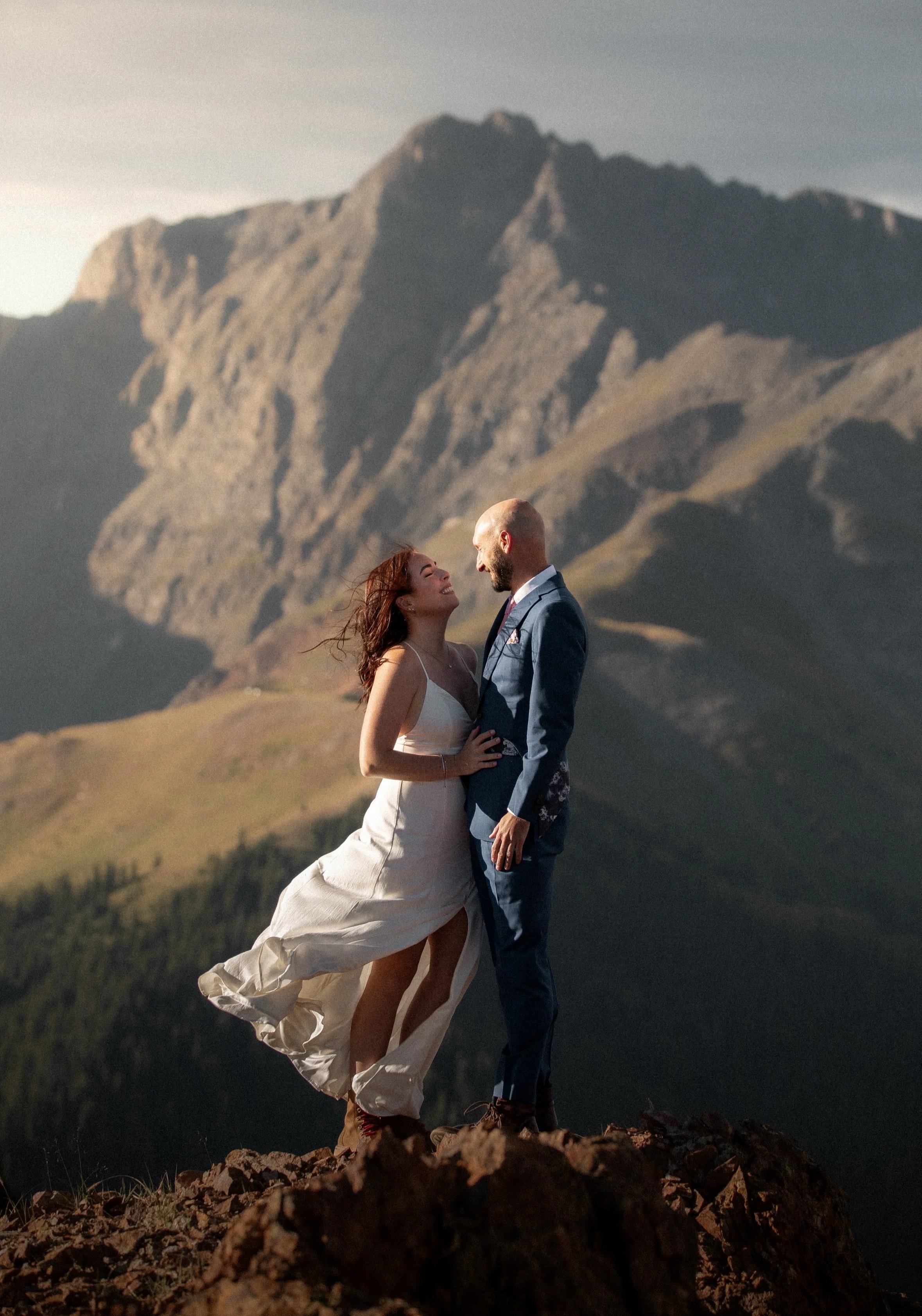 A couple, dressed in formal wedding attire, standing on a rocky ledge with a mountain landscape in the background. The woman is wearing a white dress, and the man is in a wine blue suit. They are gazing at each other, smiling.
