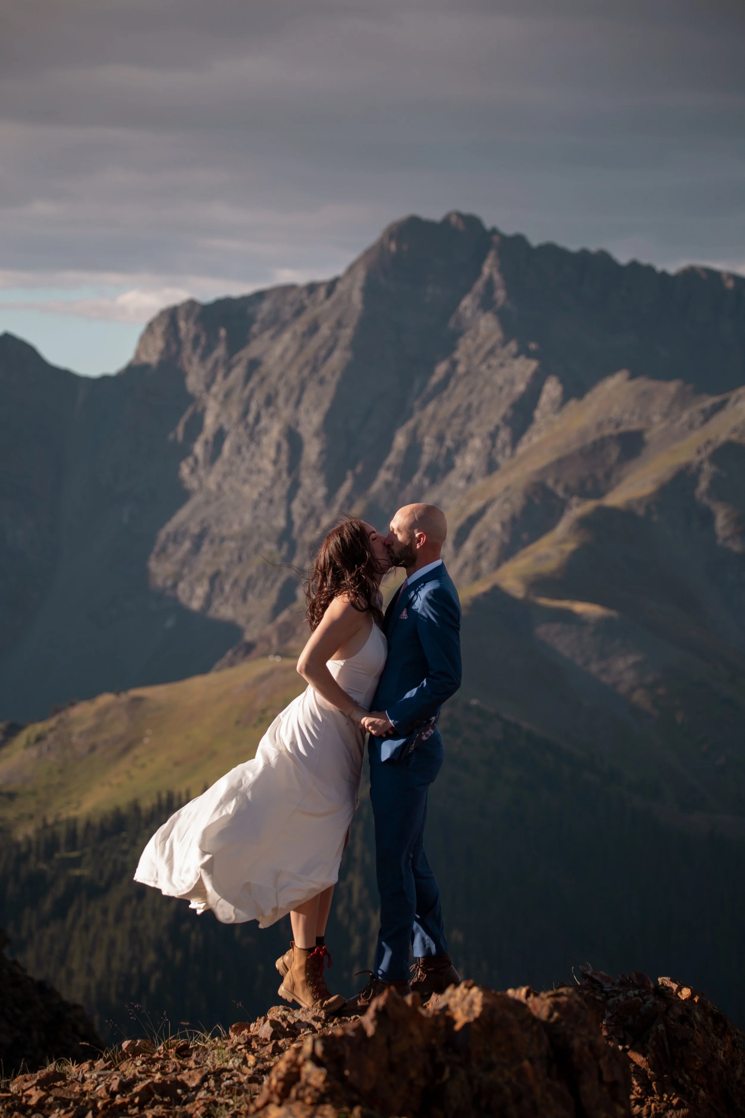 A couple dressed in wedding attire sharing a kiss on a rocky mountain ledge with a mountain range in the background.