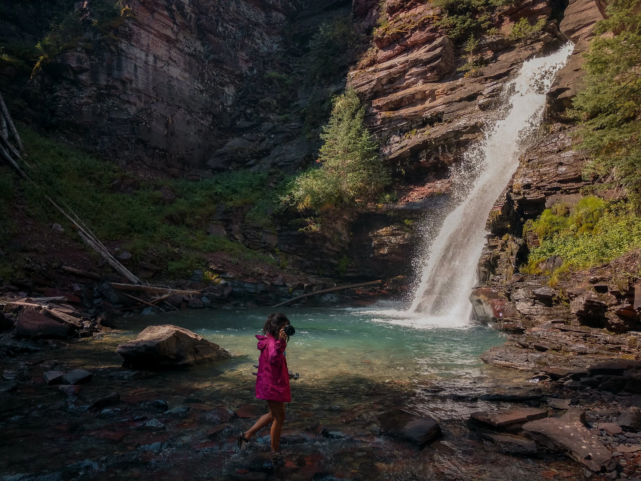 A person in a pink raincoat stands in water near a waterfall cascading down a rocky cliff in a forested area.