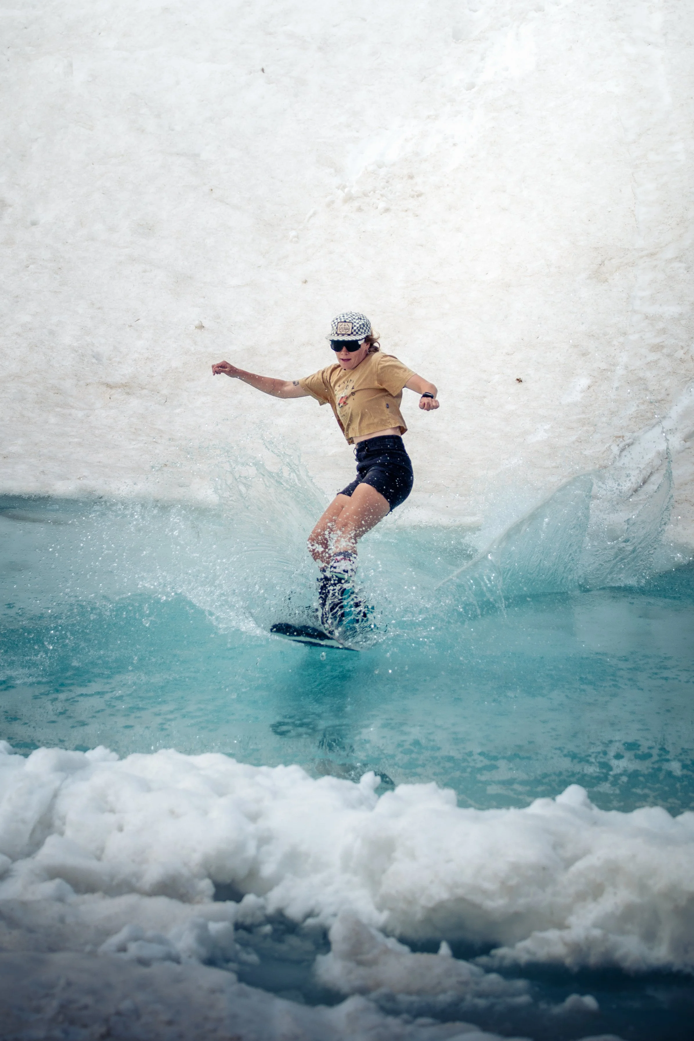 Person surfing on a wave with foam at the bottom, wearing sunglasses, a tan shirt, black shorts, and a checkered cap.
