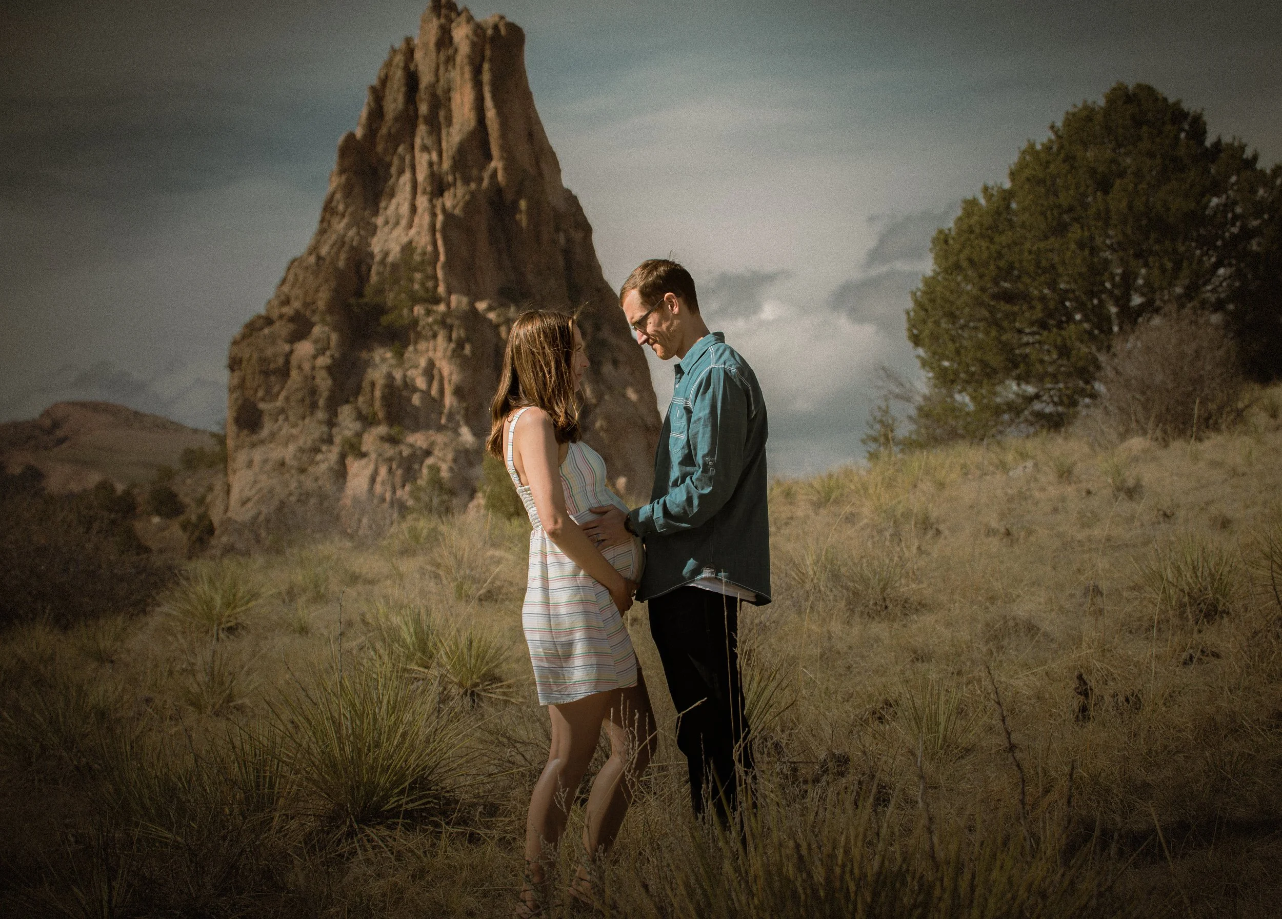 A pregnant woman and a man standing outdoors in a grassy area with a large rock formation and a tree in the background, holding hands and smiling at each other.