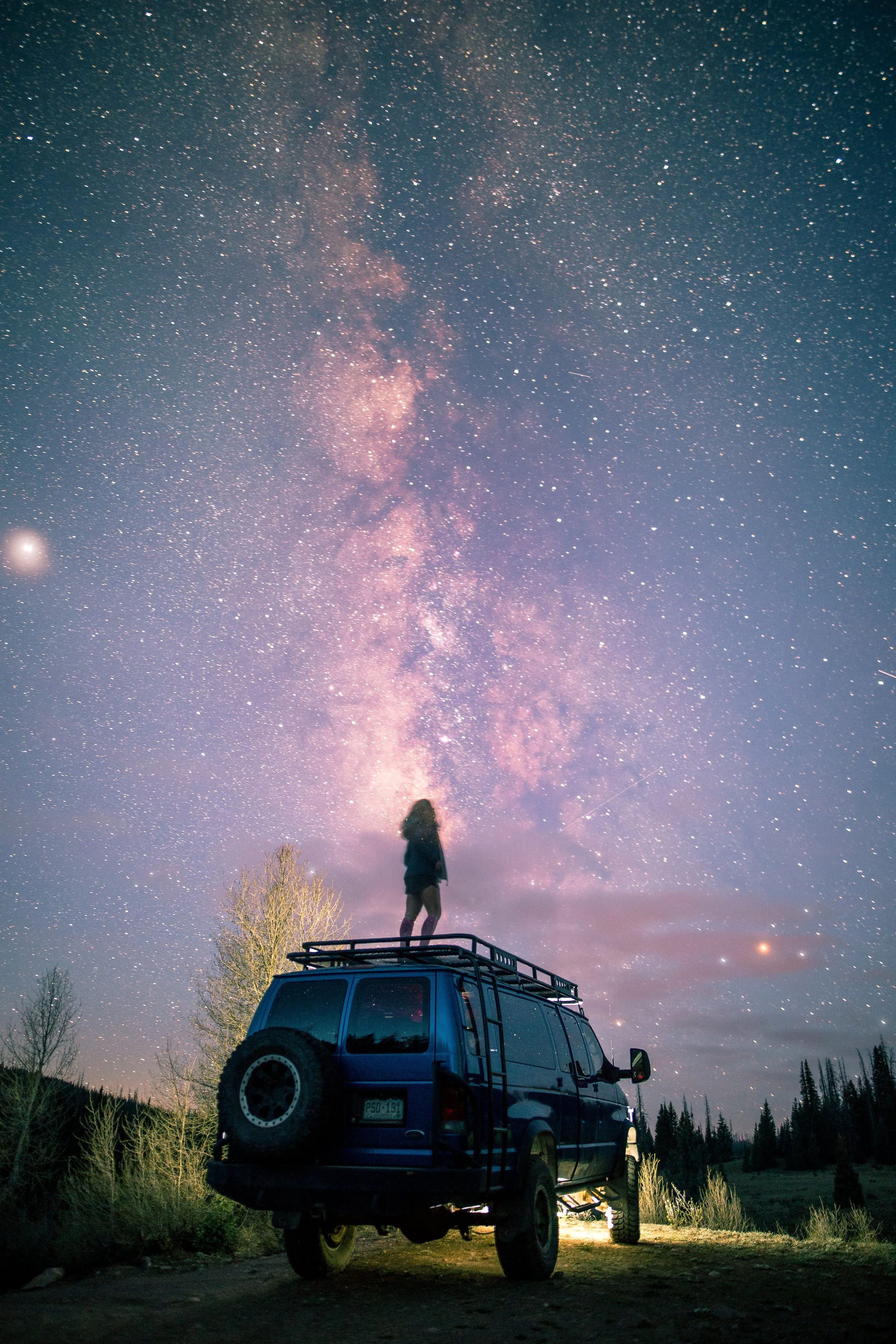 A person standing on top of a blue SUV, looking up at the starry night sky with the Milky Way galaxy visible.