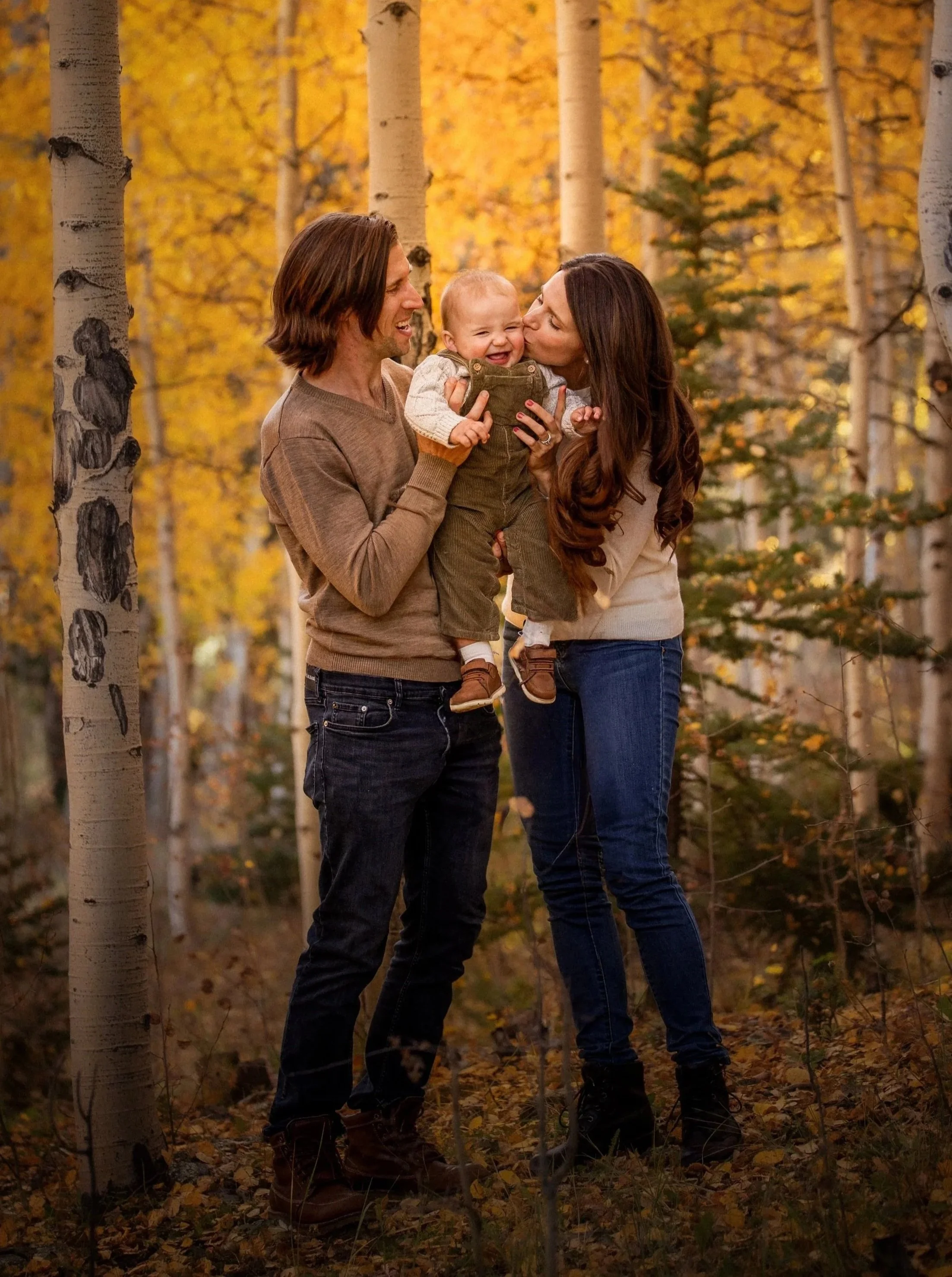 A family of three in a forest with autumn leaves, a man, woman, and a young child, smiling and playing together.