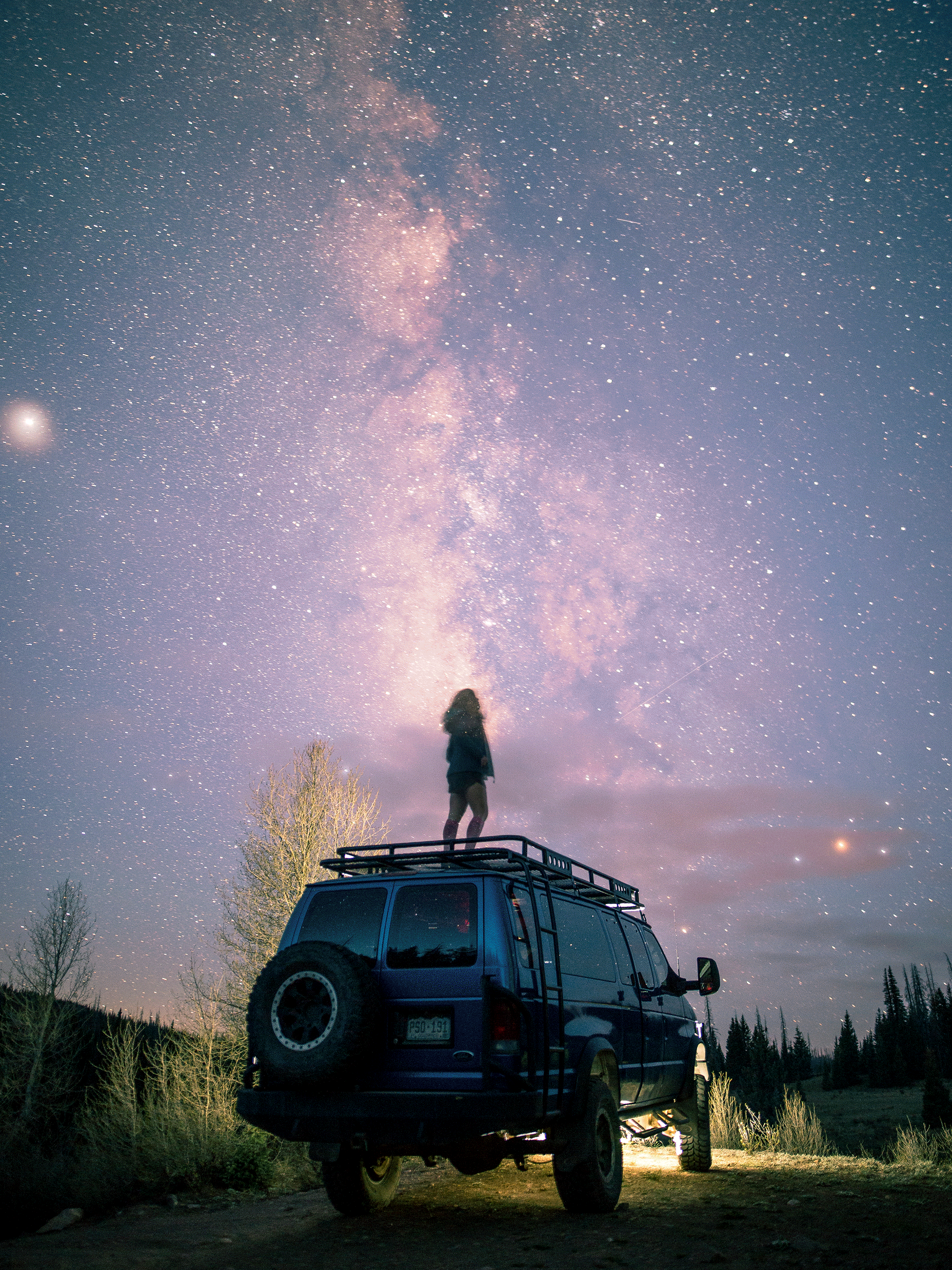 A person standing on top of a blue vehicle, gazing at the Milky Way galaxy and stars in the night sky, in a rural outdoor setting with trees and open land.