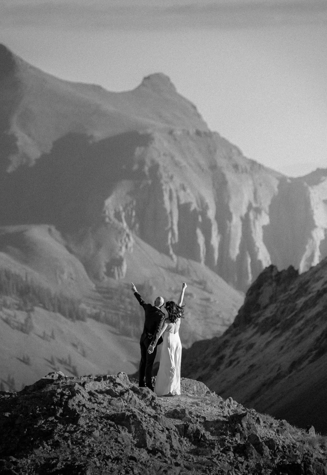 A couple in wedding attire standing on a rocky outcrop, facing a mountain range in the background, with the woman raising one arm in celebration.