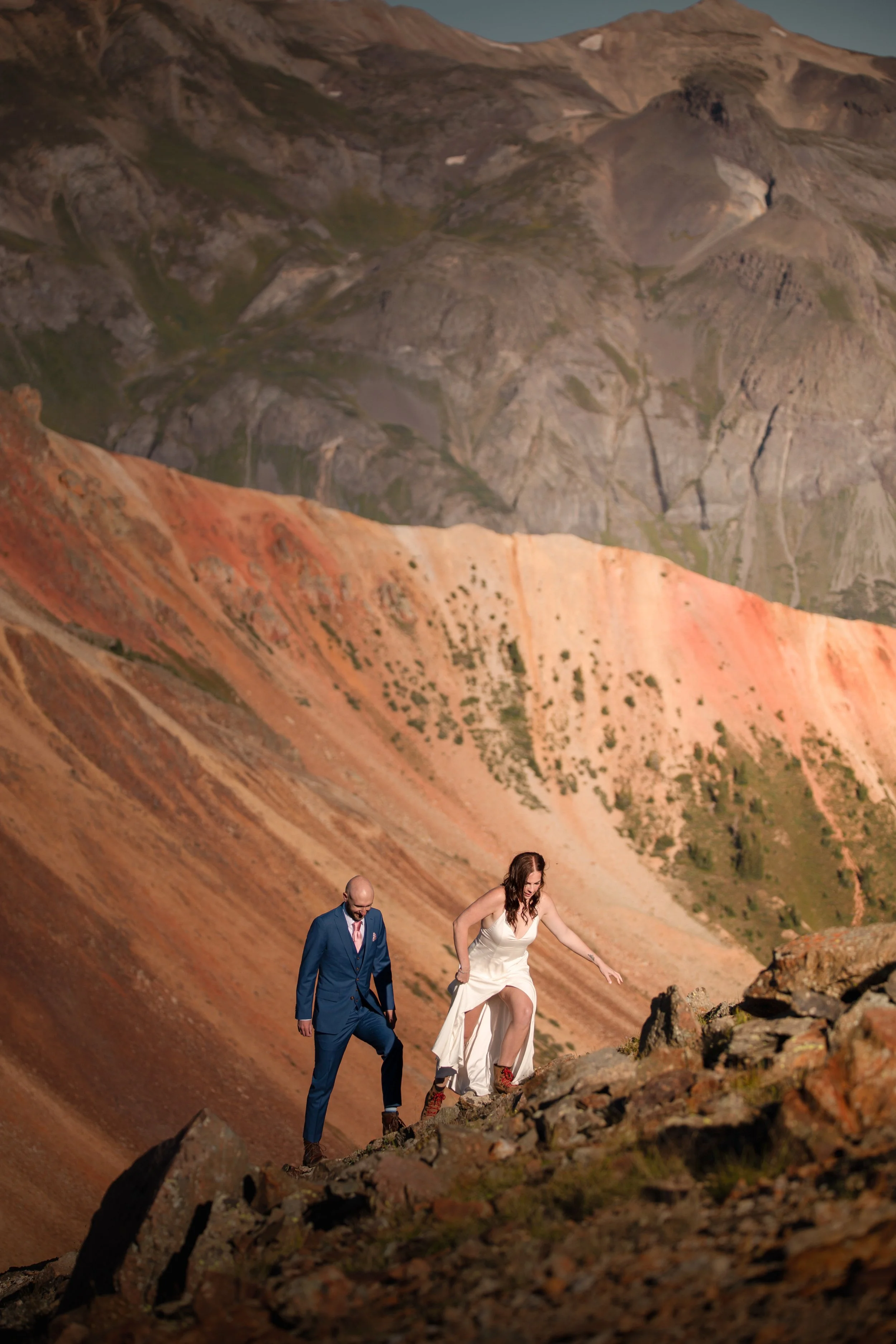 A man in a blue suit and a woman in a white dress hiking up rocky terrain on a mountain slope, with colorful mountains in the background.