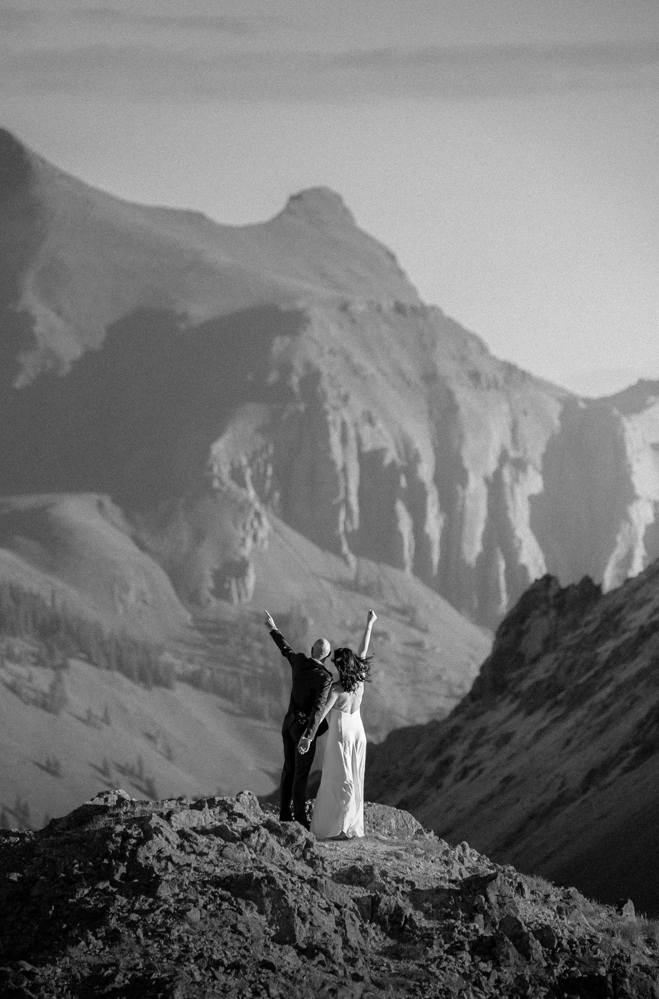 A couple stands on rocky terrain with arms raised in celebration, with mountains in the background, in a black and white photograph.