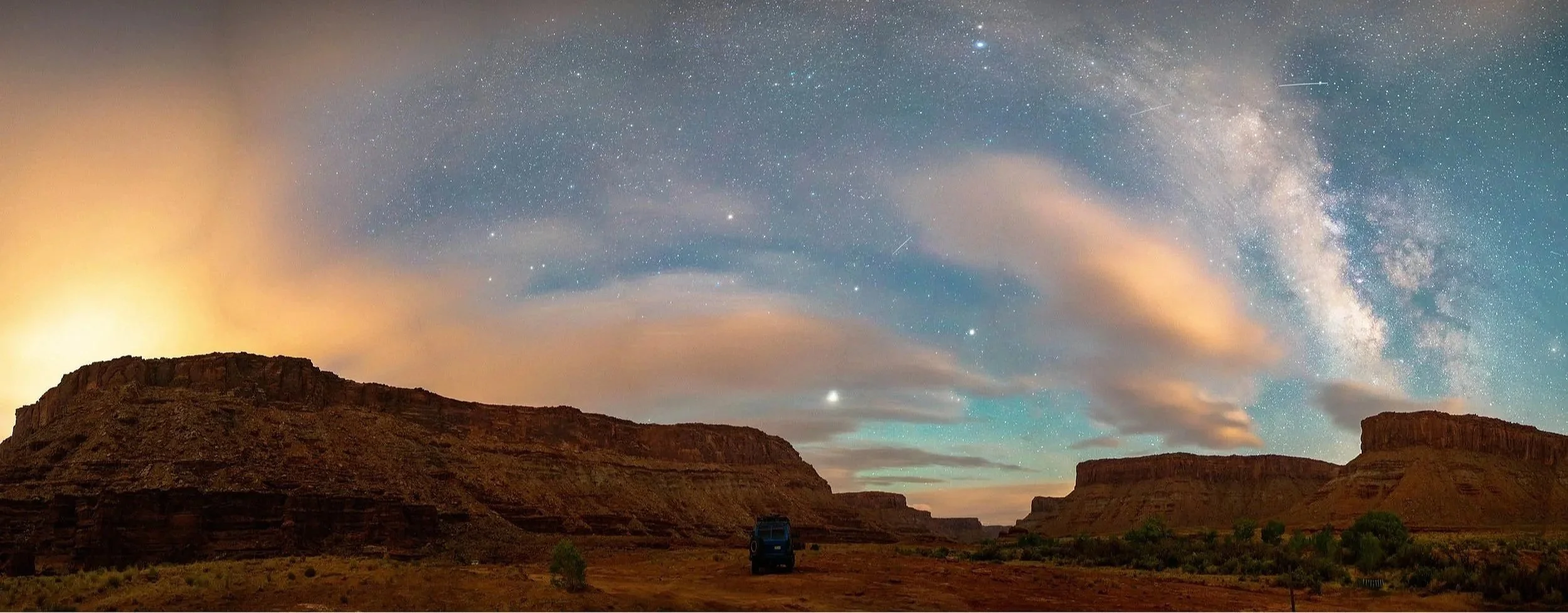 Nighttime desert landscape with canyon walls under a starry sky, including the Milky Way galaxy and shooting stars, with a vehicle parked on the ground.