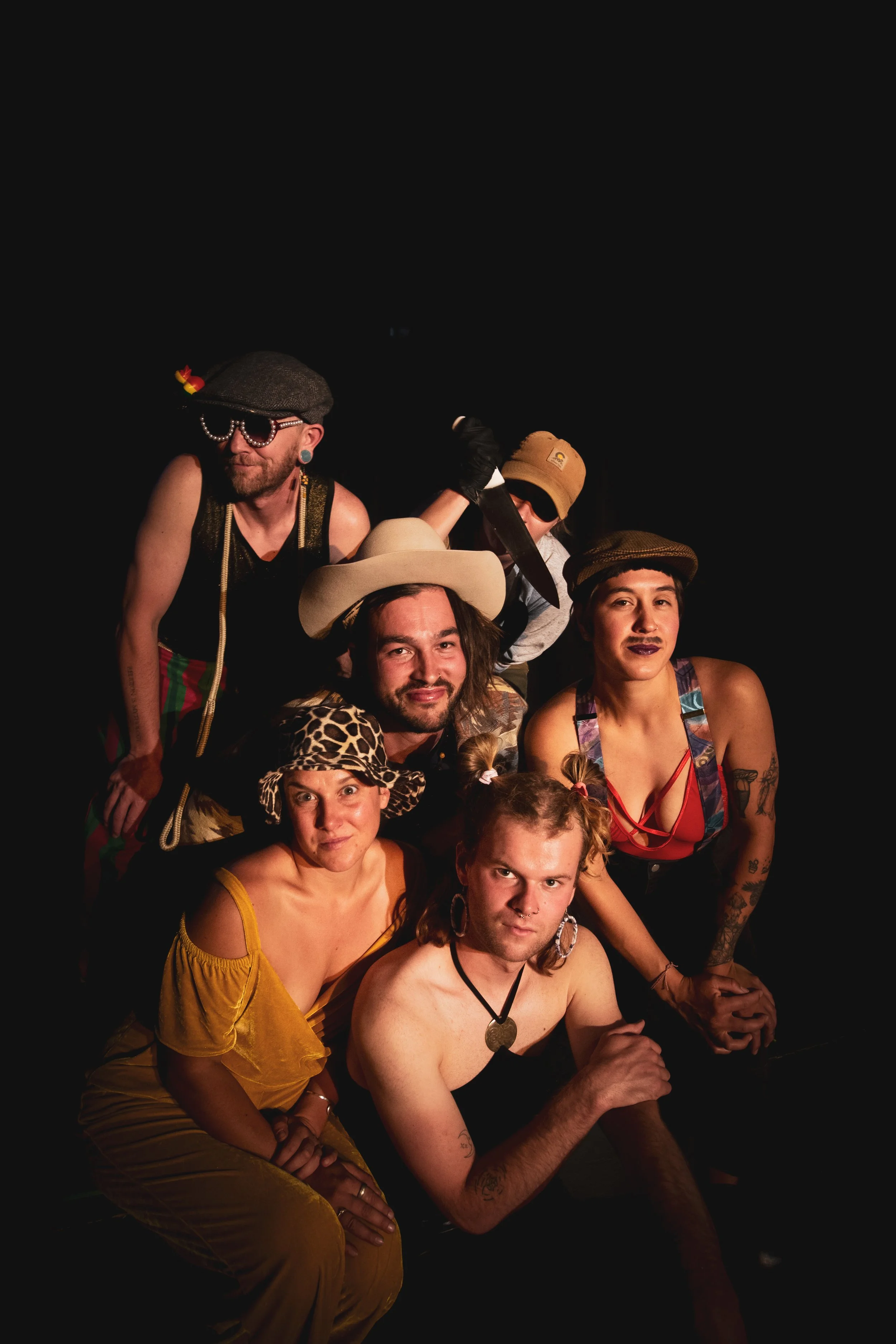 Group of six people dressed in colorful, eclectic costumes posing against a black background, with some wearing hats and makeup.