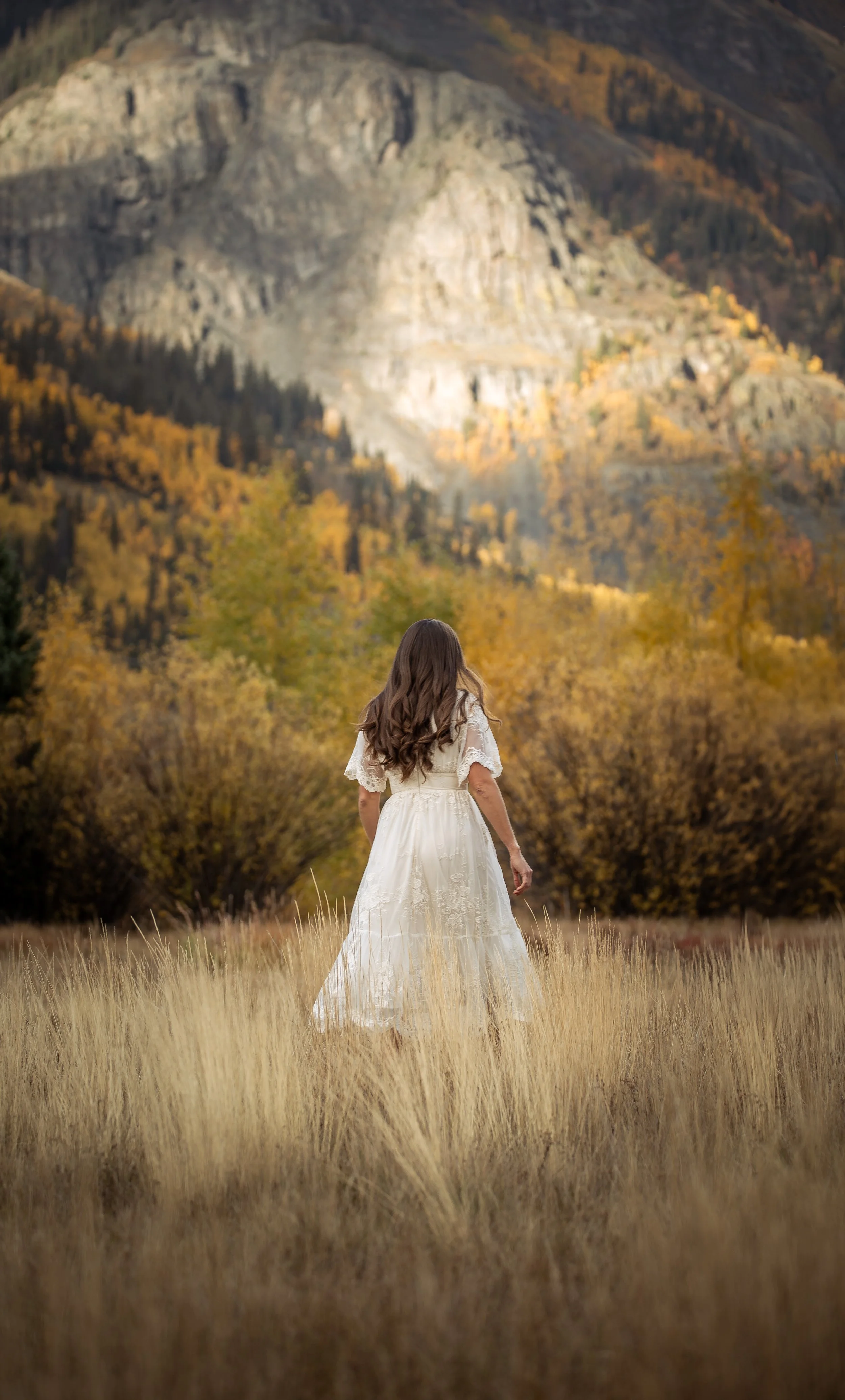 A woman in a white dress standing in a field of tall grass, facing away, with mountains and autumn trees in the background.