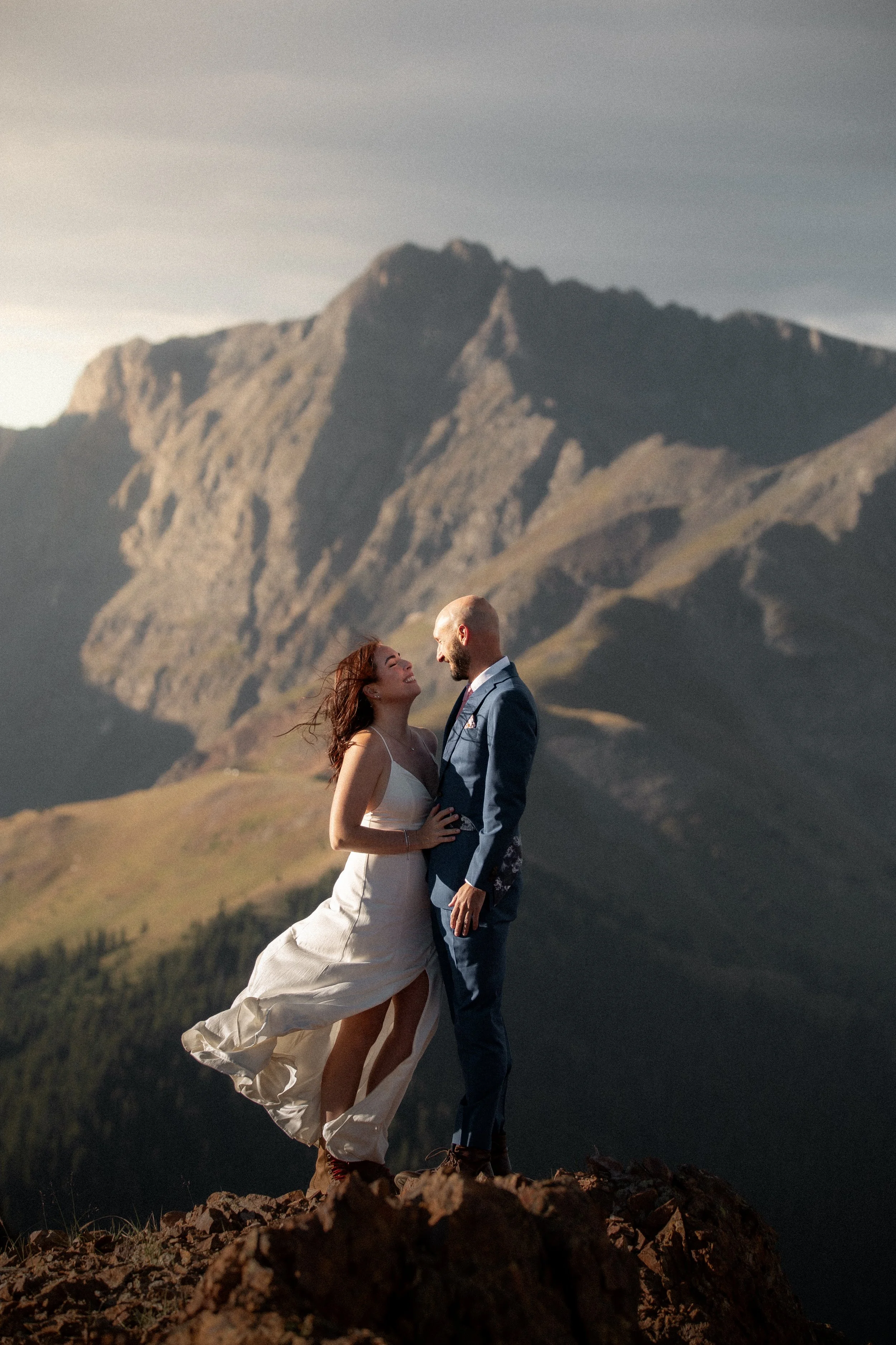 A couple in wedding attire standing close together on rocky ground outdoors with mountains in the background, smiling at each other.