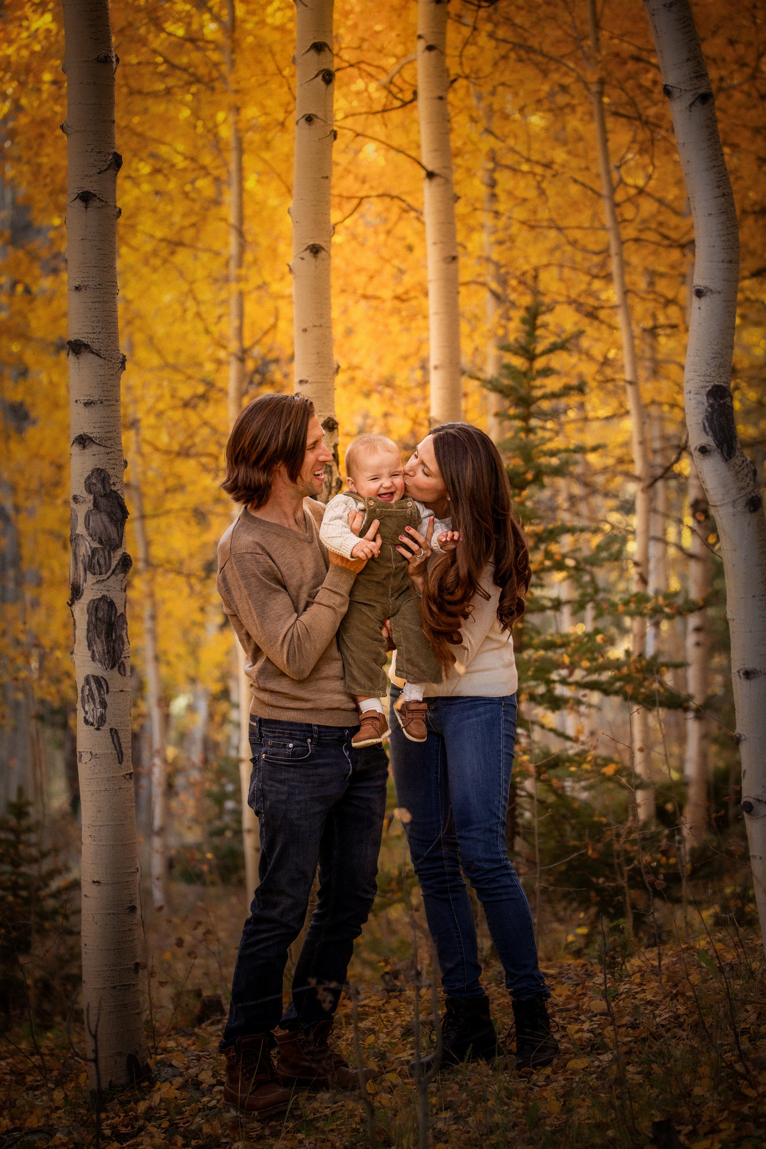 A family of three, a man and woman with a young child, enjoying time together in an autumn forest with yellow and orange leaves, standing between tall white birch trees.