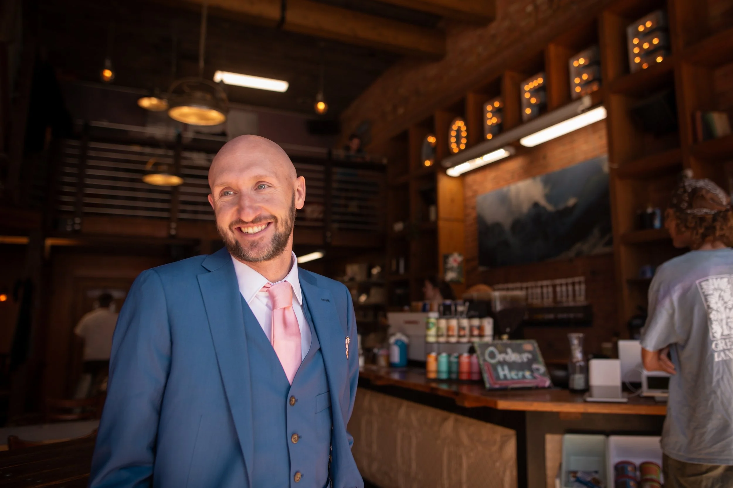 A smiling man in a blue suit and pink tie standing inside a cozy cafe or coffee shop, with a brick wall and wooden shelves in the background.
