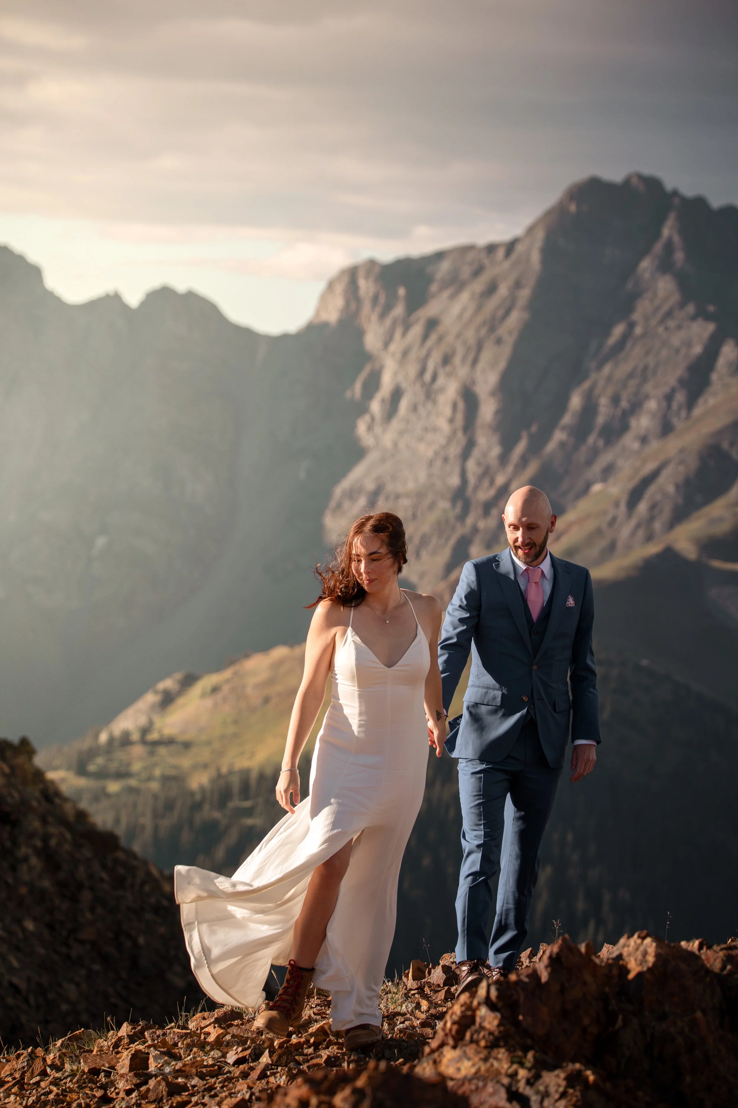 A couple, a woman in a white dress and a man in a blue suit, walking hand in hand on rocky terrain with mountains in the background.