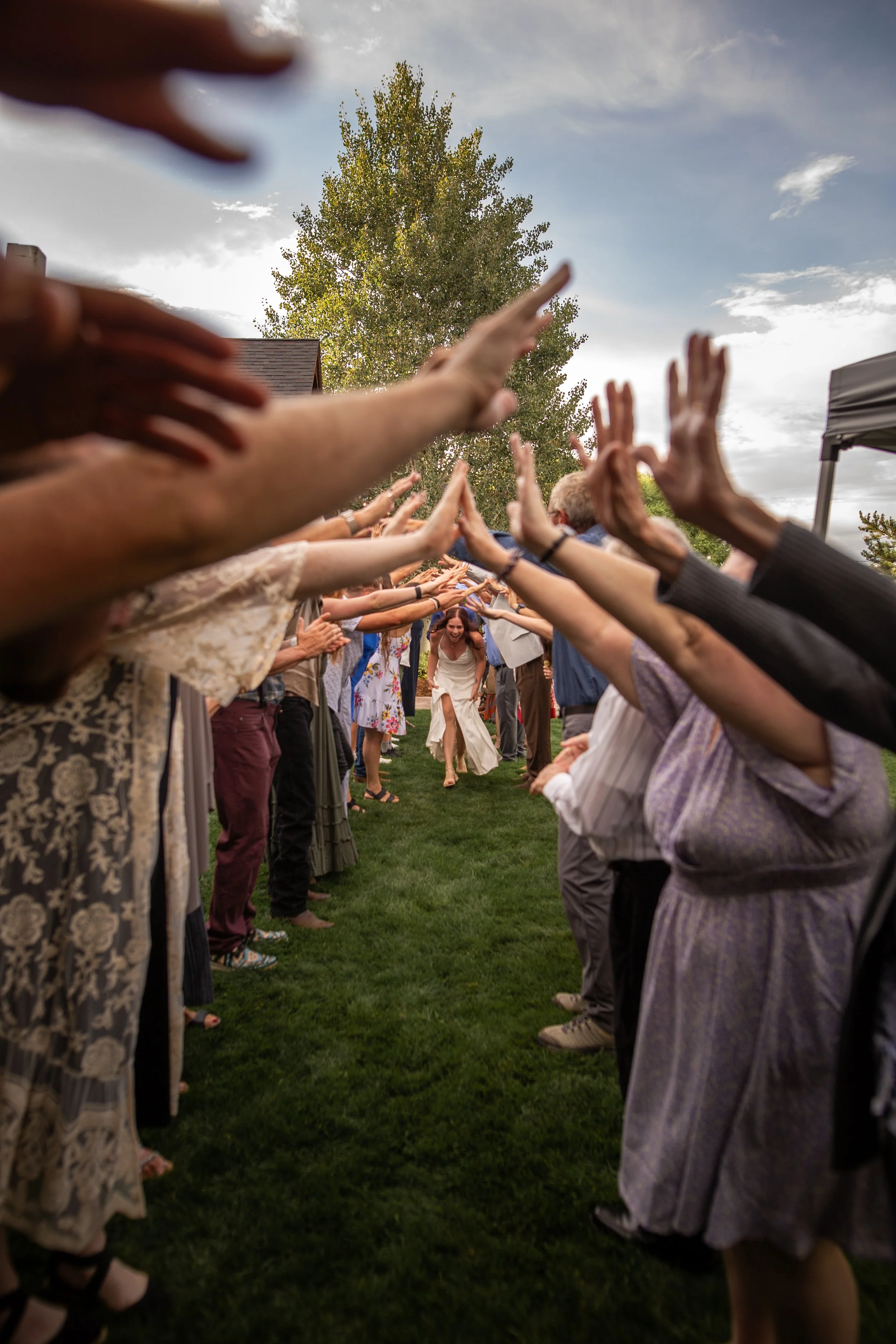 A bride walking through an outdoor archway of people with raised hands during a wedding celebration.