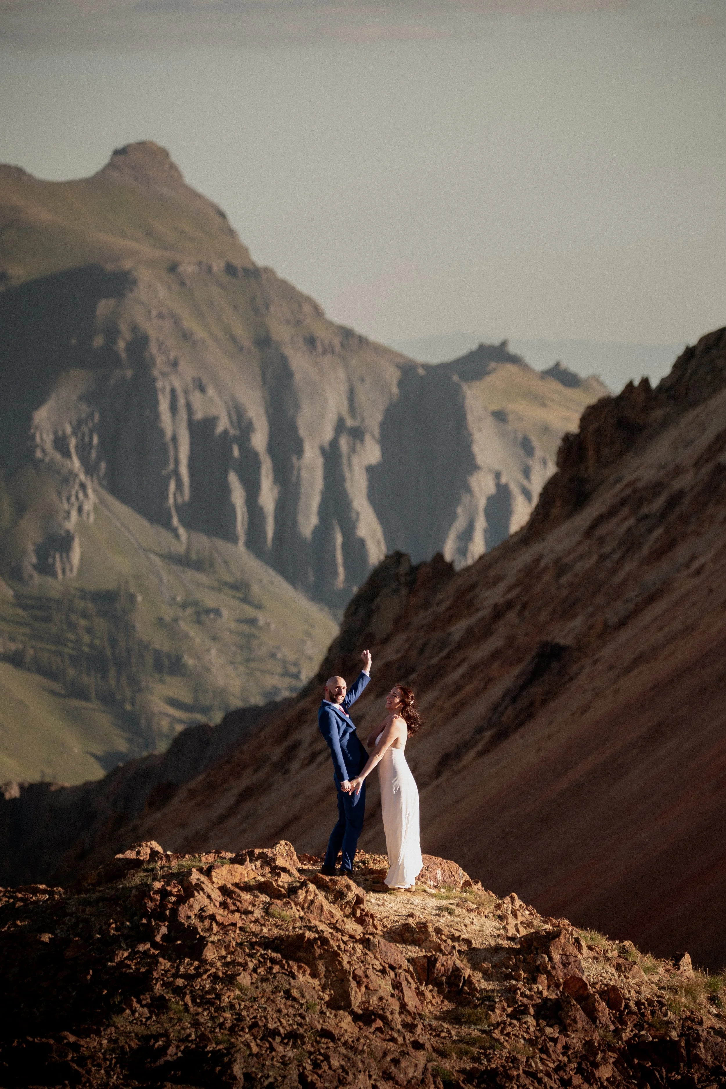 A couple in wedding attire standing on rocky terrain with a mountainous landscape in the background.