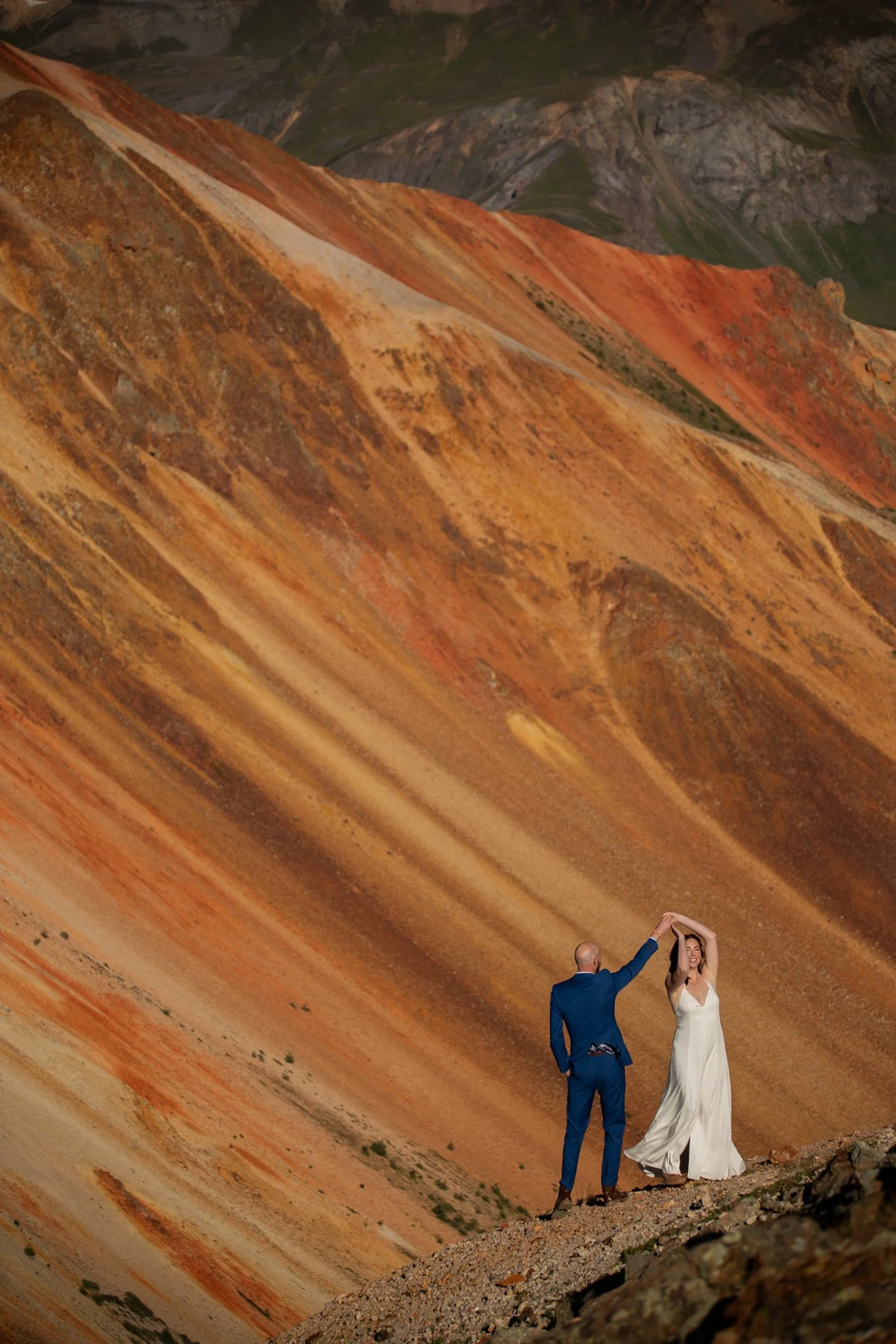 A couple, with the man in a blue suit and the woman in a white dress, standing on rocky terrain at the base of a colorful mountain with orange, red, and green hues, during daylight.