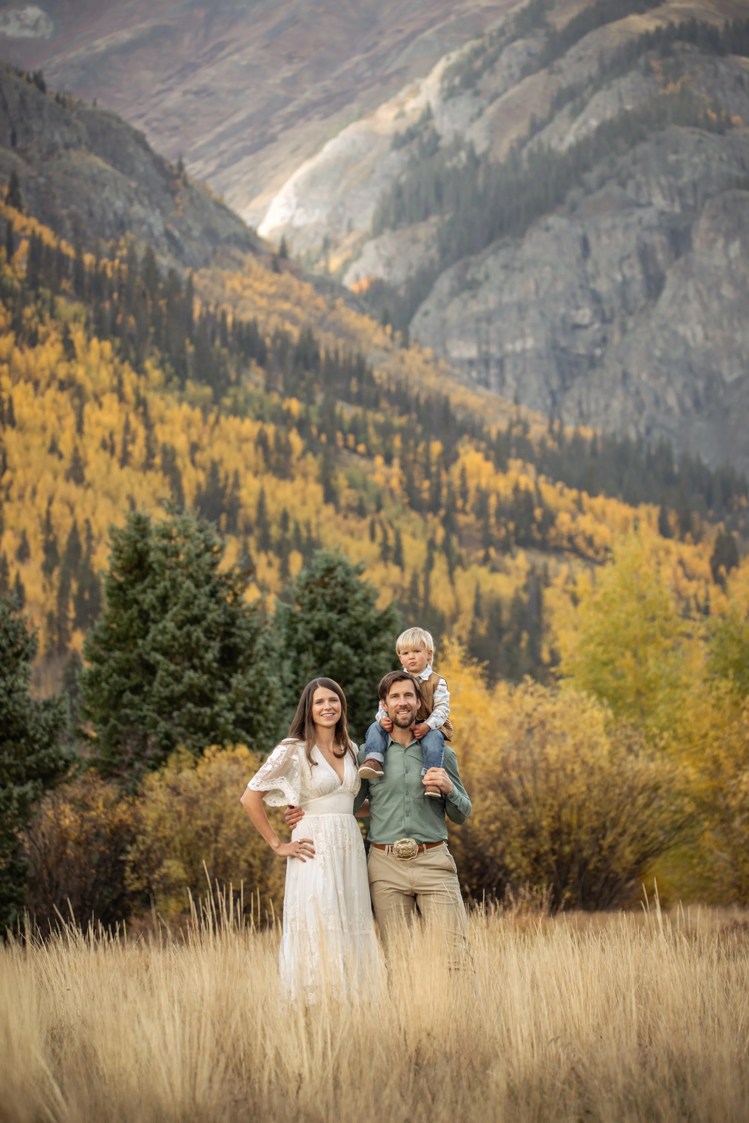 A family of three enjoying a walk in a field surrounded by trees and mountains during fall. The woman is wearing a white dress, the man is in a green shirt with khakis, and a young boy is sitting on the man's shoulders.