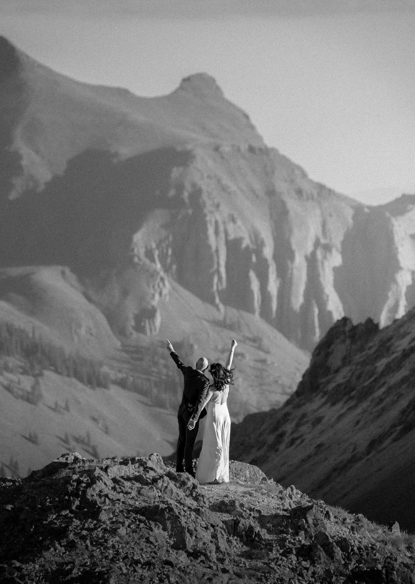A black-and-white photo of a couple dressed formally, standing on a rocky hilltop with arms raised, against a backdrop of large cliffs and mountainous landscape.