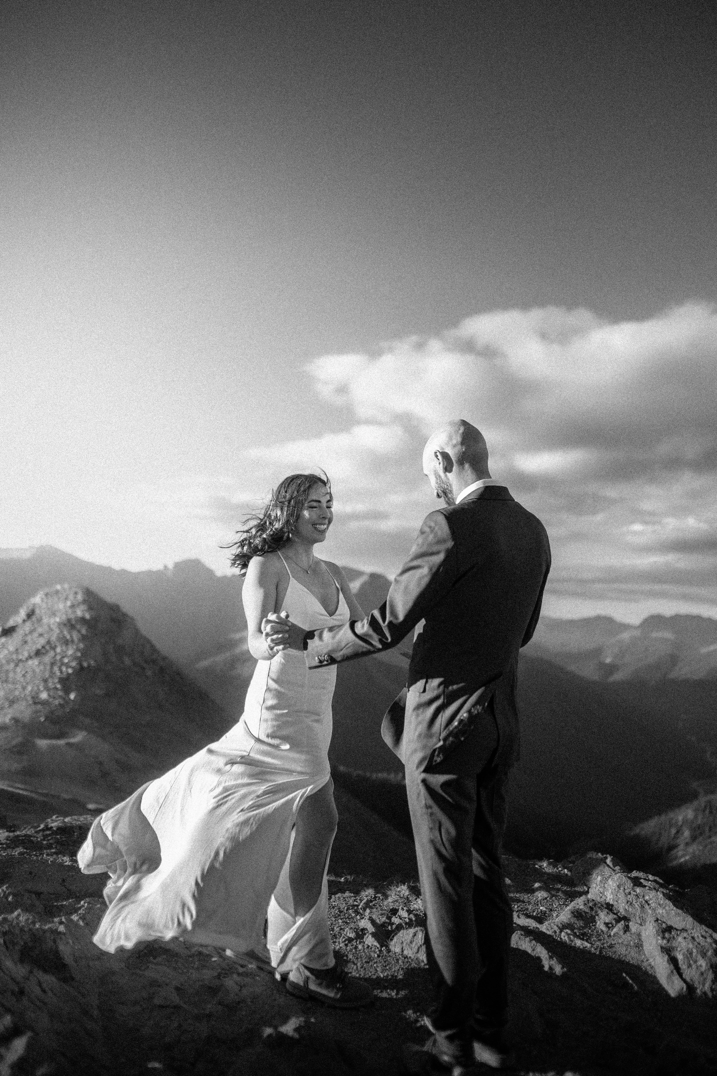 A black and white photo of a couple holding hands and dancing on a mountain top, with a mountainous landscape and clouds in the background.