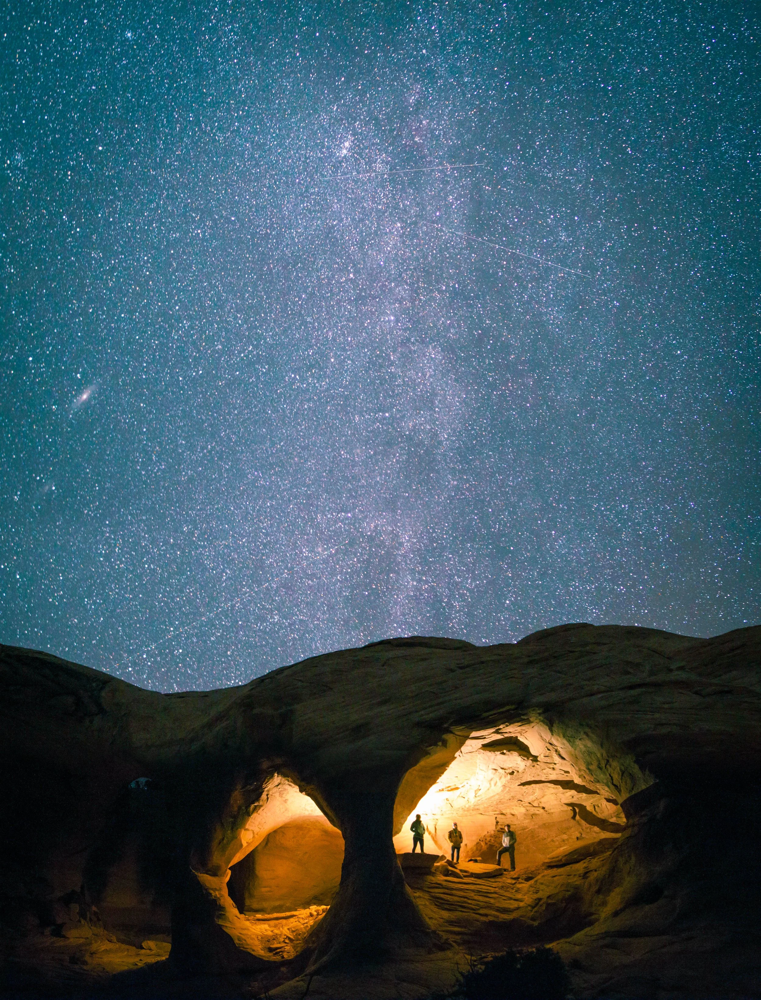 Group of three people inside a natural rock formation at night, illuminated by warm light, with a starry sky and the Milky Way galaxy overhead.