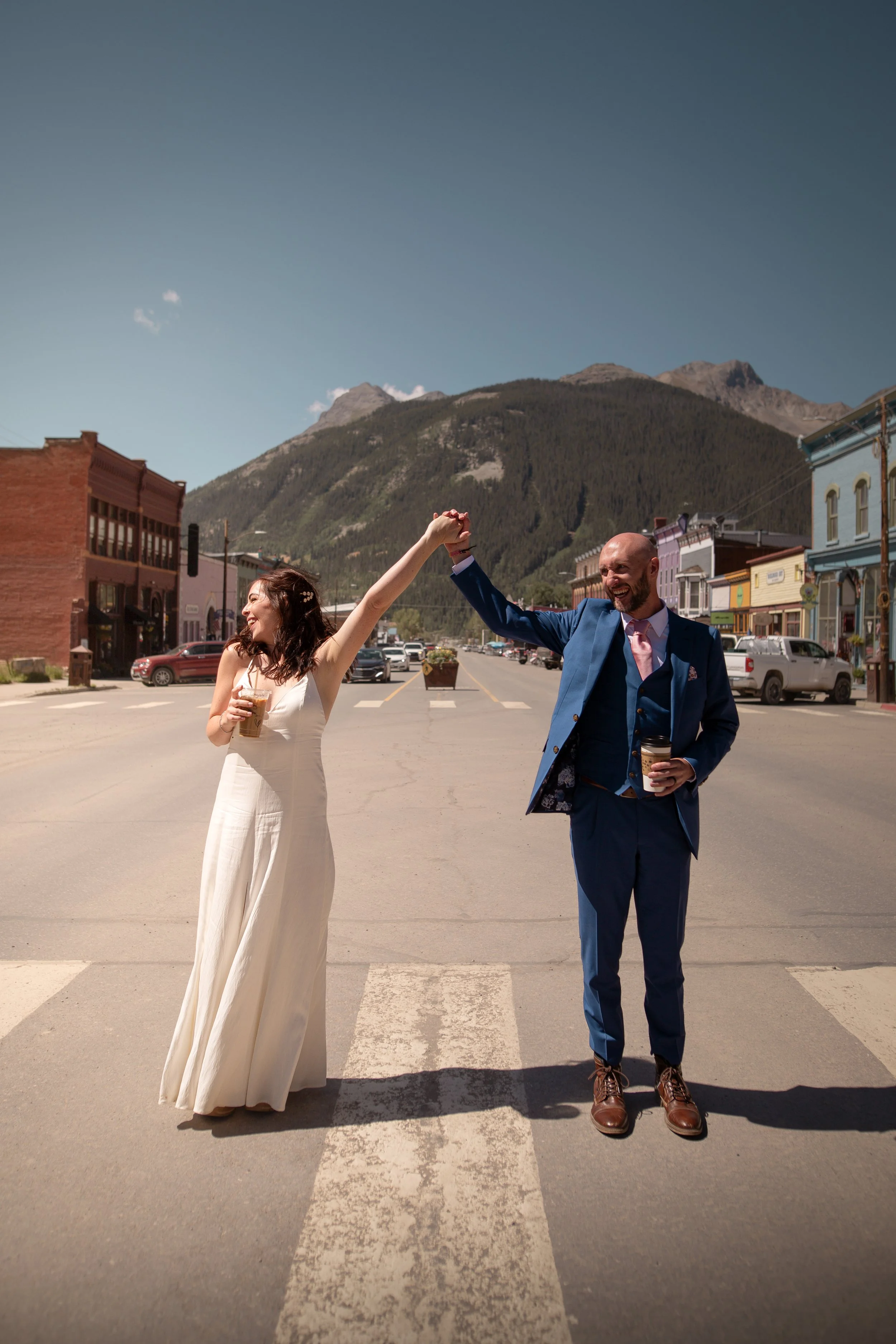 A newlywed couple dancing on a crosswalk in a small town, with mountains in the background, holding coffee cups and smiling.