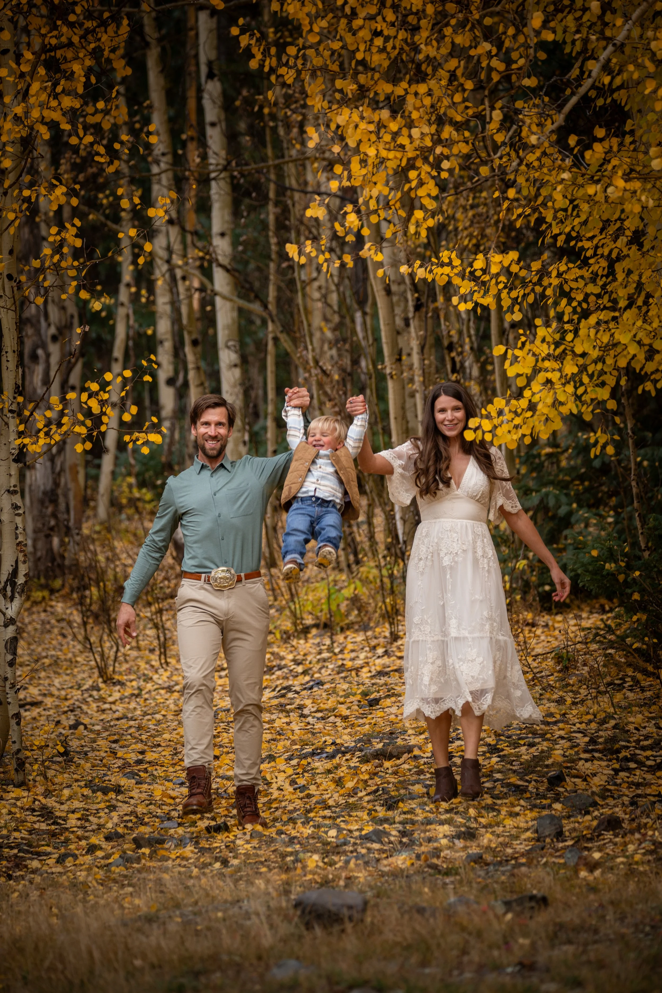 A family of three walking through a forest in autumn, with the father and mother holding their young son between them. The father is holding the boy's hand, lifting him up, and all three are smiling. The forest is filled with yellow leaves, and some 