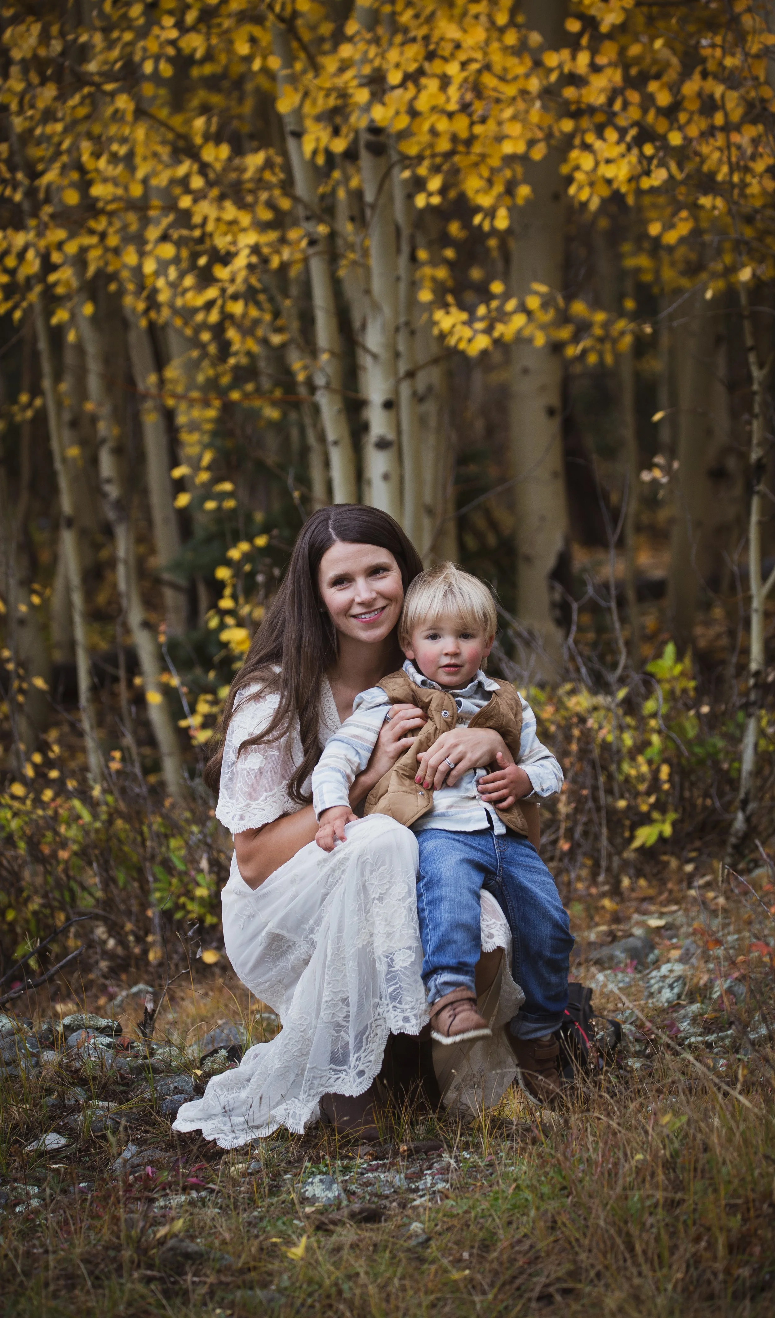 A woman and young boy sitting outdoors in a forest with autumn yellow leaves. The woman has long brown hair and is wearing a white lace dress. The boy has blonde hair and is dressed in blue jeans, a striped shirt, and a brown vest.