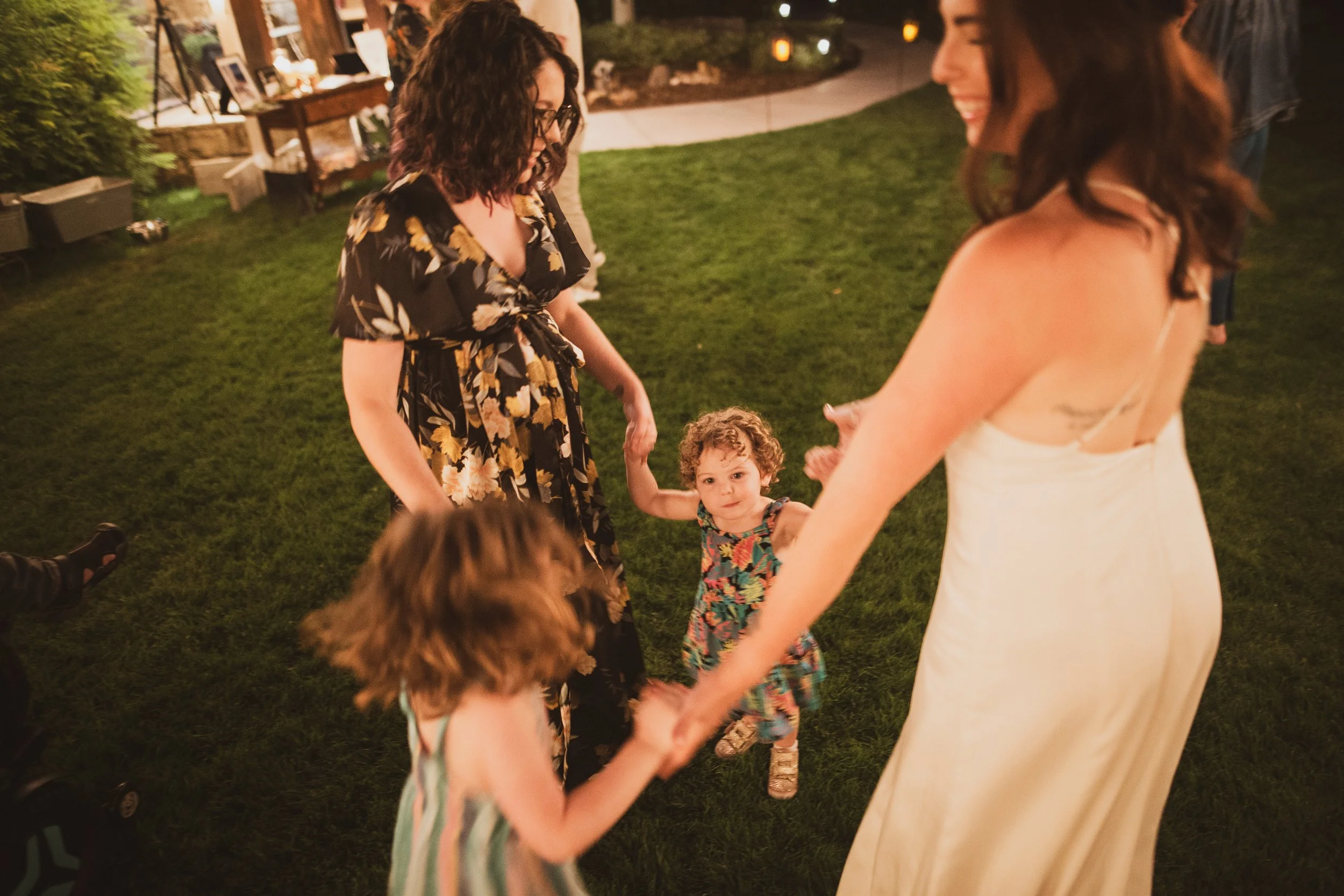 Two adult women are holding hands with two young girls on a grassy lawn at night, smiling and dancing together.