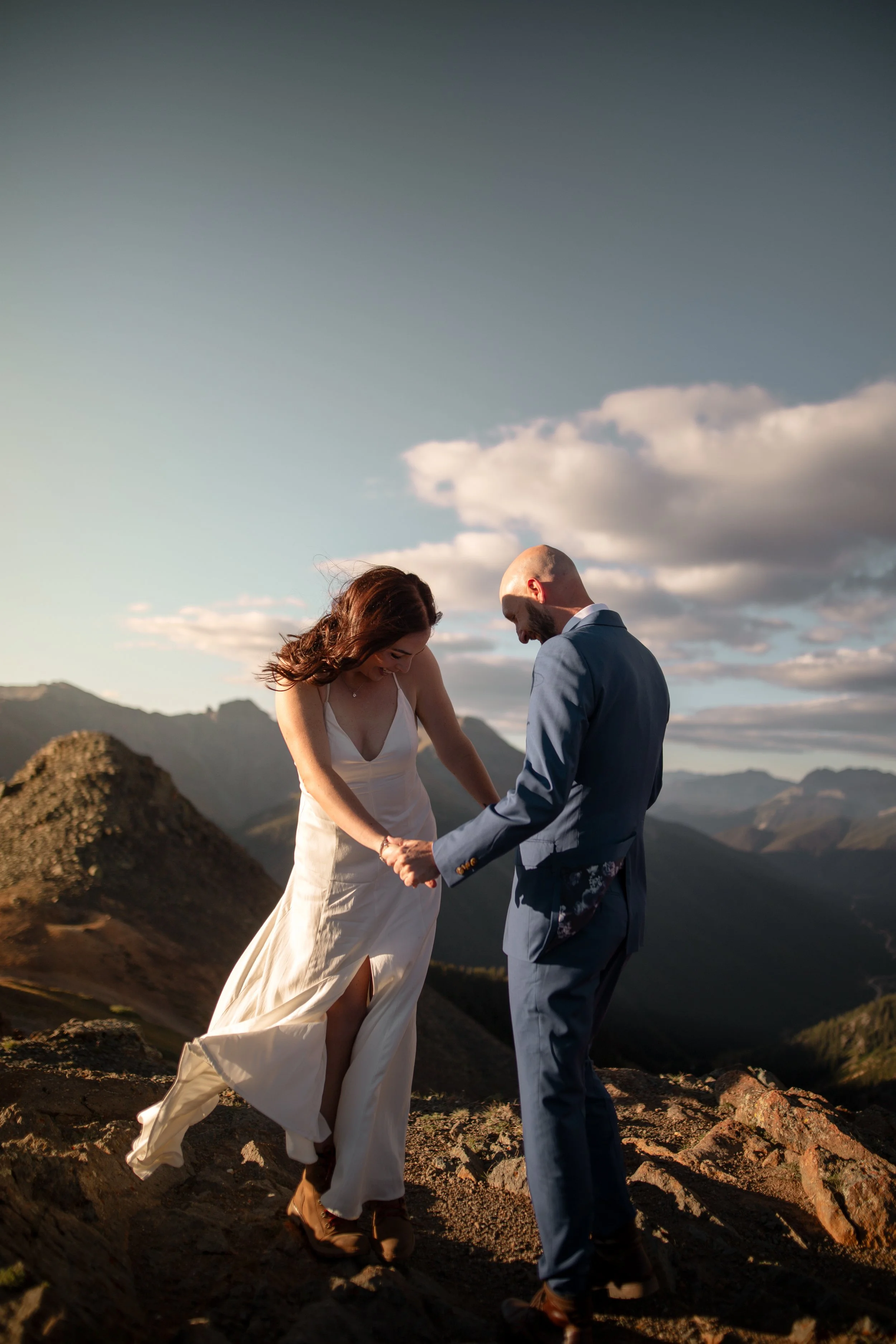 A couple in wedding attire holding hands and laughing outdoors on a mountain landscape